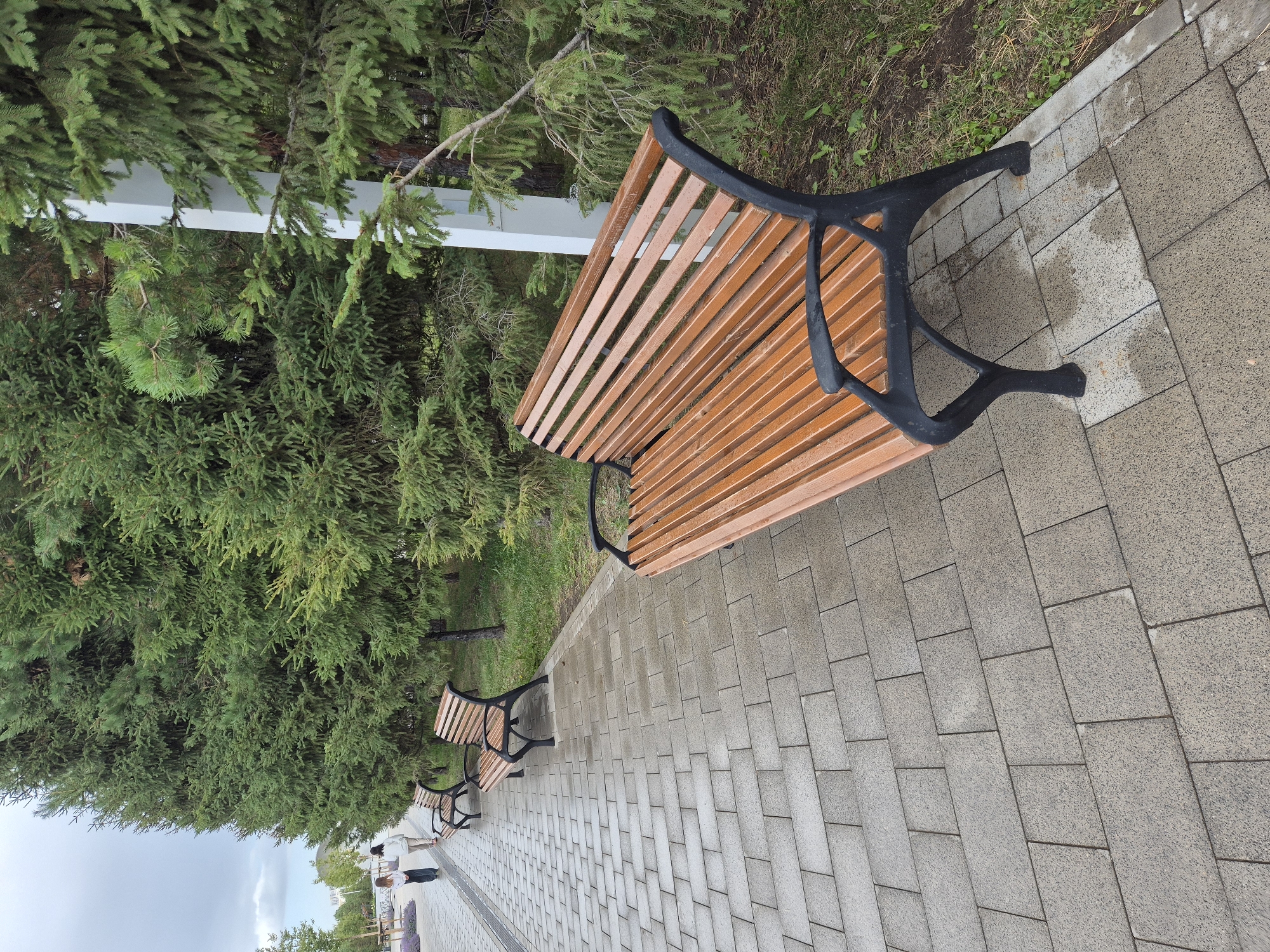 The image depicts a park scene in Astana, Kazakhstan, featuring a paved walkway lined with mature coniferous trees. Several wooden park benches with black metal frames are placed along the walkway. In the background, two individuals, likely women, are walking away from the camera. The sky is overcast, suggesting a cloudy day. The ground appears to be wet, possibly from recent rain. A white vertical post, possibly a lamp post or signpost, is visible amongst the trees. The foreground is dominated by the texture of the paving stones and a close-up view of a park bench.