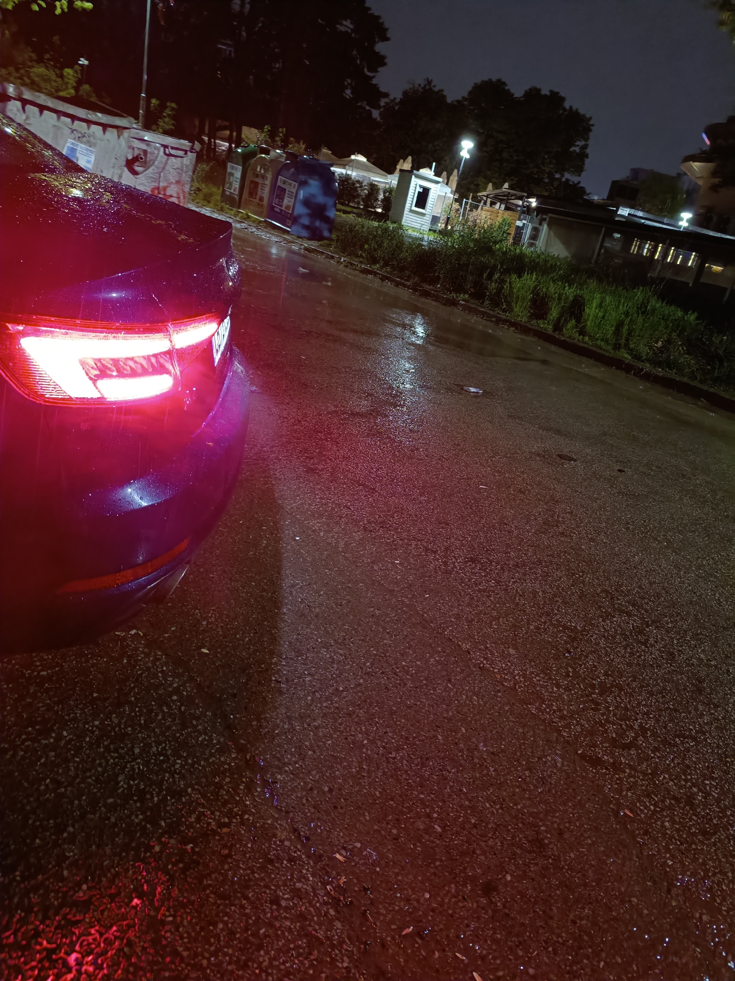 This nocturnal image captures a wet street scene, likely in Sofia, Bulgaria, as indicated by the "EVROPAK" recycling bin featuring text "За по-чиста София" (For a cleaner Sofia). The main subject is the rear left quarter of a dark-colored car, possibly black or dark blue, with its bright red taillights prominently illuminated. Water droplets cling to the car's surface, and the asphalt road is visibly wet and reflective, suggesting recent rain or an ongoing drizzle.
In the background on the left, several large waste containers are visible, including grey communal dumpsters and two blue recycling bins. One blue bin clearly displays "EVROPAK" and the aforementioned Bulgarian text. Beyond these, trees and dimly lit building structures emerge from the darkness. To the right of the road, a strip of overgrown grass and vegetation separates the street from more buildings and outdoor structures further back, some with lit windows or streetlights. The scene is devoid of people, conveying a quiet, reflective mood typical of a rainy night in an urban or suburban area. The streetlights and car lights cast glows and reflections on the wet surfaces, adding to the atmospheric quality.