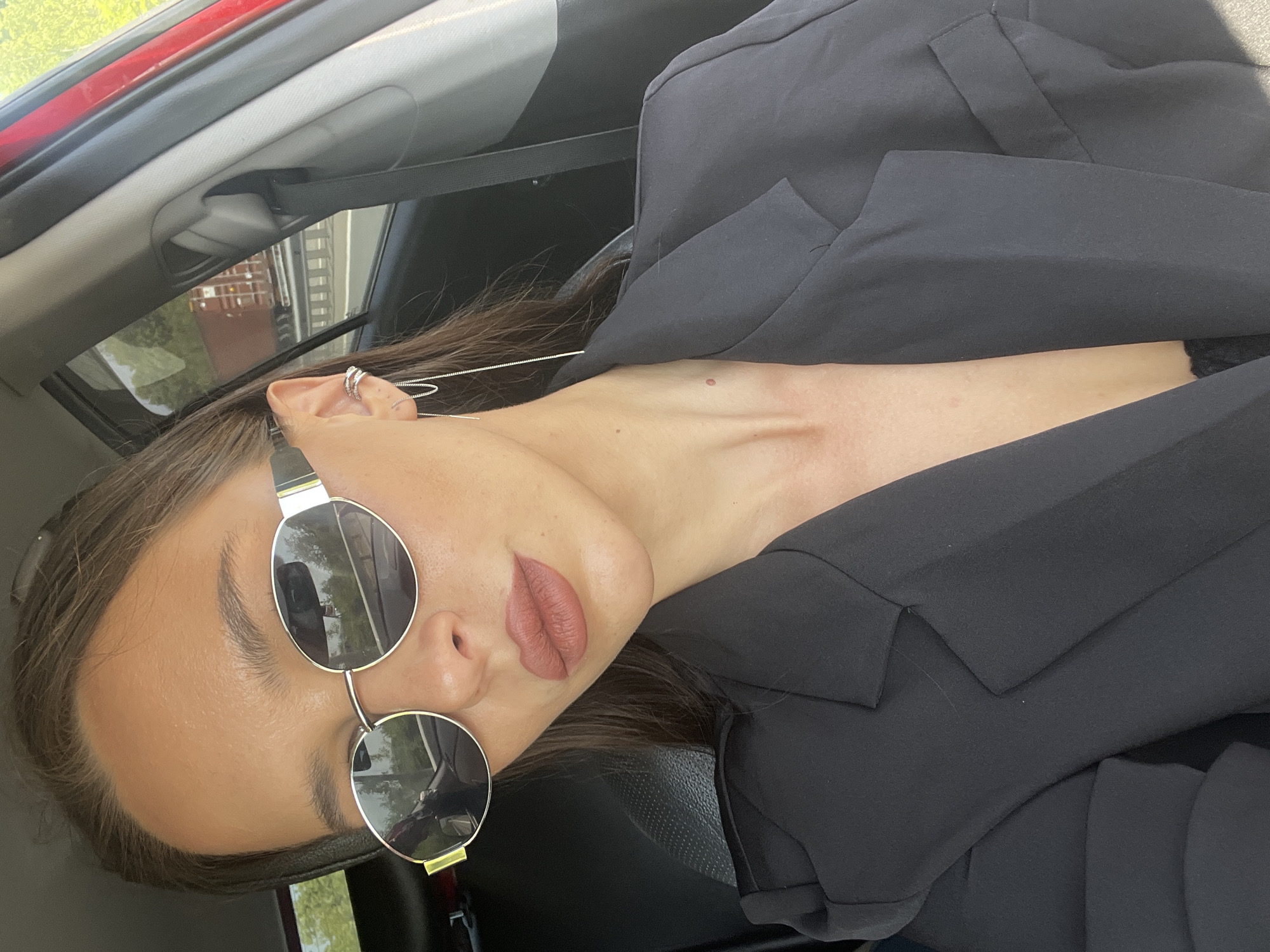 The image is a close-up, portrait-style photograph, rotated 90 degrees counter-clockwise, showing a person from the chest up inside a car. The individual, who appears to be female, is the primary subject, seen wearing large, round sunglasses with reflective lenses and a black blazer or jacket. She has dark hair, styled down, and her lips are painted with a muted, neutral-toned lipstick. Her visible right ear is adorned with multiple small ear cuffs and a long, delicate chain earring that dangles down.
The setting is the interior of a car, with parts of the headliner, a seat, and a red door frame visible in the background. Bright daylight streams in, creating highlights on the person's skin and strong reflections in the sunglasses. These reflections offer a glimpse of the outside environment, showing trees or foliage and what appears to be structures or buildings, suggesting a sunny outdoor day. The person is looking directly towards the camera, seemingly posing for a photograph. The overall impression is casual and stylish. There is no visible text in the image. This scene is set in De-Friz, Russia.