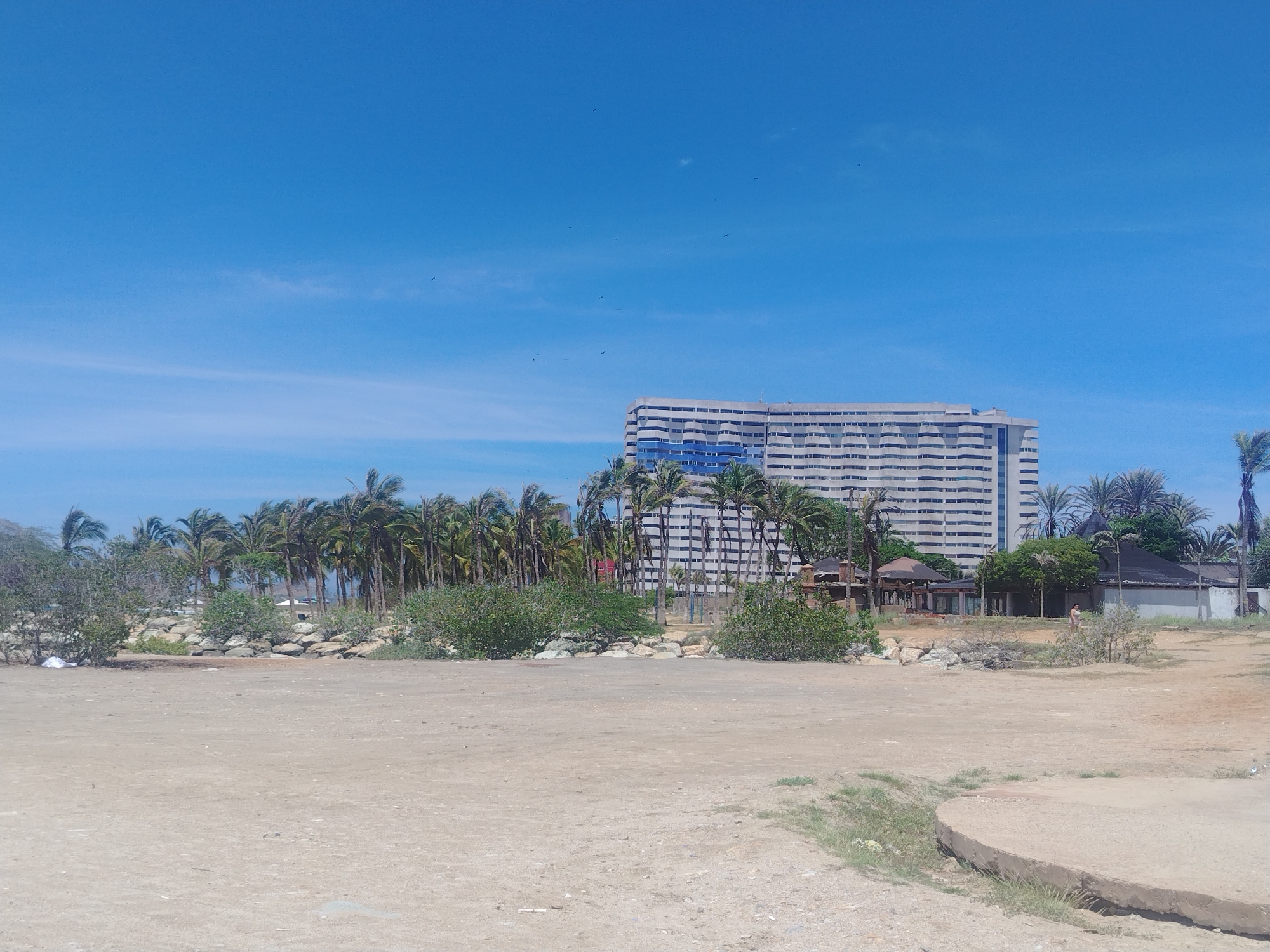 This media file captures a sun-drenched scene in Porlamar, Venezuela. The foreground is dominated by a sandy, flat expanse, with a concrete curb partially visible on the right. Beyond the sandy area, a line of lush green trees and palm trees forms a natural barrier, with scattered rocks interspersed.
In the middle ground, a large, modern building with a striking blue and white facade stands prominently. This multi-story structure appears to be a hotel or resort, evidenced by its numerous windows and balconies. Adjacent to the resort, several smaller buildings with thatched roofs and open-air structures suggest amenities like restaurants or recreational areas. A single person is visible walking in this area, adding a sense of scale and human presence.
The background is a clear, vibrant blue sky, dotted with a few faint wisps of clouds and several small, dark specks that appear to be birds in flight. The bright sunlight and clear sky indicate that it is daytime, likely mid-morning to late afternoon, with fair weather. The presence of palm trees and the architectural style of the resort strongly suggest a tropical or subtropical coastal location, consistent with Porlamar. The overall mood is one of a tranquil, sunny day at a resort destination. No visible text is present in the image.