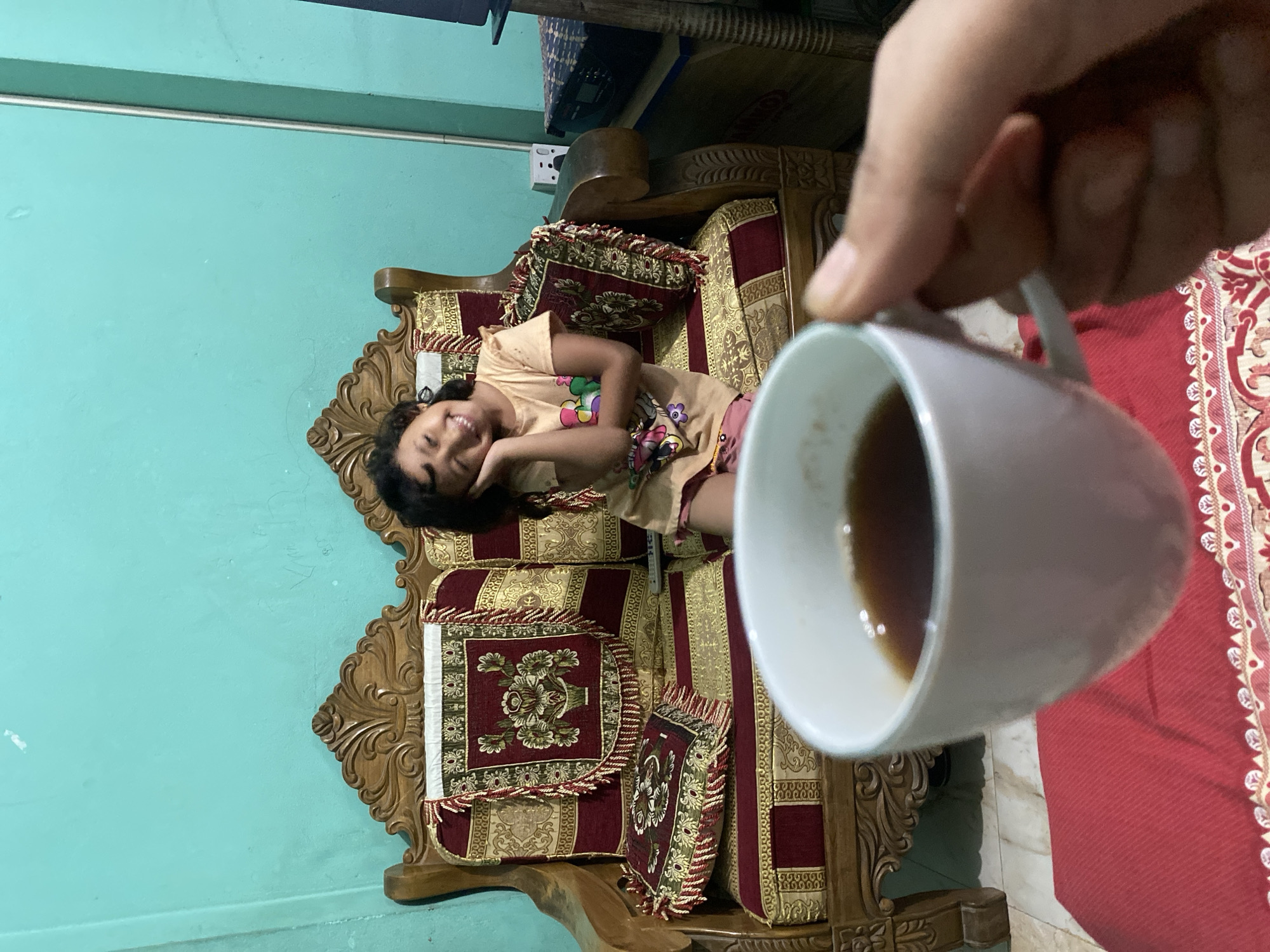 This image captures a warm, casual indoor scene, likely within a home in Dhaka, Bangladesh. The primary subject is a young girl, who appears to be a child, lying relaxed on an ornate wooden sofa. She has dark, curly hair and is smiling broadly at the camera, conveying happiness and contentment. She is dressed in a light-colored, short-sleeved top adorned with vibrant, colorful patterns, possibly cartoon characters or floral designs, and matching patterned shorts or trousers.
The sofa is a prominent piece of furniture, featuring intricately carved wooden frames and plush cushions upholstered in a rich red fabric with elaborate gold patterns and decorative fringes. Several cushions and a smaller decorative pillow are arranged on the sofa, enhancing its inviting appearance.
In the blurred foreground, a person's hand is visible, holding a white ceramic mug containing a dark liquid, likely coffee or tea. The background features a plain teal-colored wall, creating a lively backdrop. Above the sofa, some dark, indistinct objects, possibly part of another piece of furniture or electronics, are visible, along with a white electrical outlet. A section of light-colored tiled flooring is seen to the right of the sofa, with a red patterned rug or carpet partially visible further right.
The image exudes a relaxed and informal atmosphere, portraying a moment of everyday life and the joyful expression of the child. No discernible text is visible in the frame.