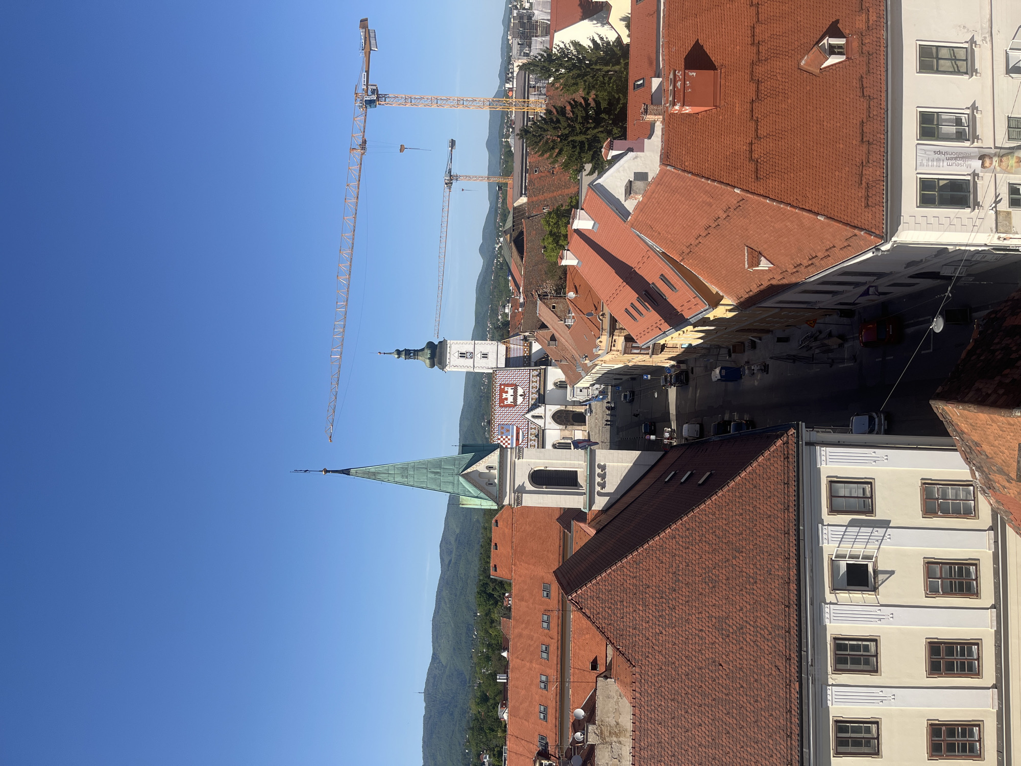 This is a daytime aerial shot of Zagreb, Croatia. The view encompasses a town square, complete with its prominent St. Mark's Church. The church's green spire and the distinctive Croatian coat-of-arms on its roof are focal points. Red-tiled roofs of adjacent buildings dominate the foreground. In the distance, a mountain range marks the horizon. Cranes punctuate the skyline, suggesting construction or urban development. Below, a road has vehicles.
