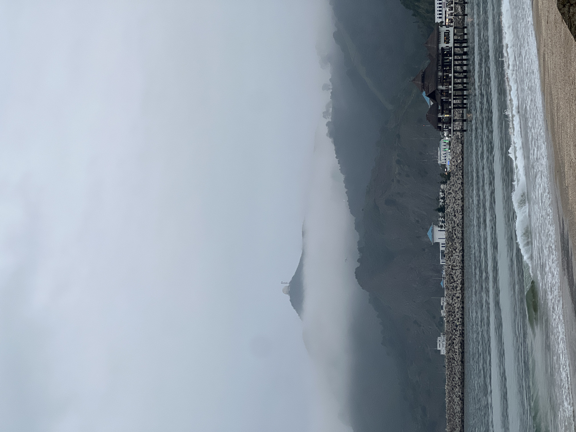 The image captures a misty coastal landscape in M'Diq, Morocco, with a dominant mountain range partially obscured by dense fog or low-lying clouds. In the distance, a distinct white, cylindrical structure, possibly a lighthouse or monument, can be seen emerging from the mist on a mountain peak.
Along the base of the mountains, a coastline features a sandy beach and a long breakwater constructed from numerous concrete tetrapods. Behind this barrier, a series of buildings line the shore. Some structures have distinctive bright blue roofs, while others are larger, multi-level complexes with dark brown, tiered roofs, possibly resorts or hotels, some appearing to extend over the water on stilts or platforms. Minimal artificial light is visible from these buildings.
The foreground shows the ocean with white-capped waves gently breaking onto the sandy shore. The water appears calm closer to the breakwater. The heavy fog creates a soft, ethereal, and somewhat subdued atmosphere, limiting visibility. The time of day appears to be overcast or early morning/late afternoon due to the diffused light. No people or specific activities are visible, and there is no discernible text in the image.