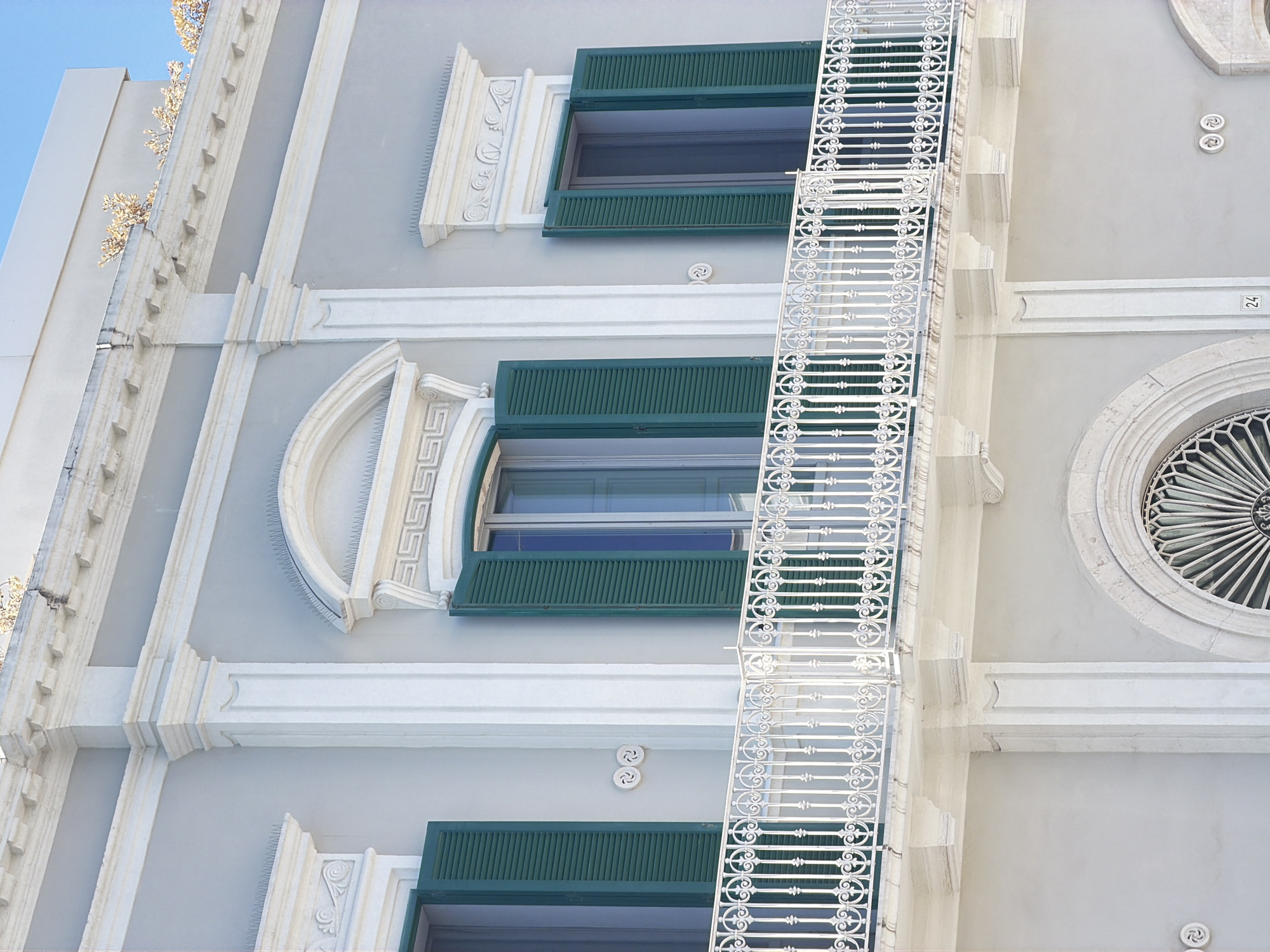 The image shows a close-up view of a building facade with neoclassical architectural elements in Polignano a Mare, Italy. The building is painted a light grey, and the facade features white decorative moldings and columns. There are three windows visible on the upper floor, each with dark green shutters that are partially open. The central window is arched, while the two side windows are rectangular. A white wrought-iron balcony with an intricate scrolling pattern runs across the facade in front of these windows. Below the balcony, the building has another level with a decorative circular window and a number "24" visible on the wall. The sky above is a clear blue. The scene is static, with no people or animals present.