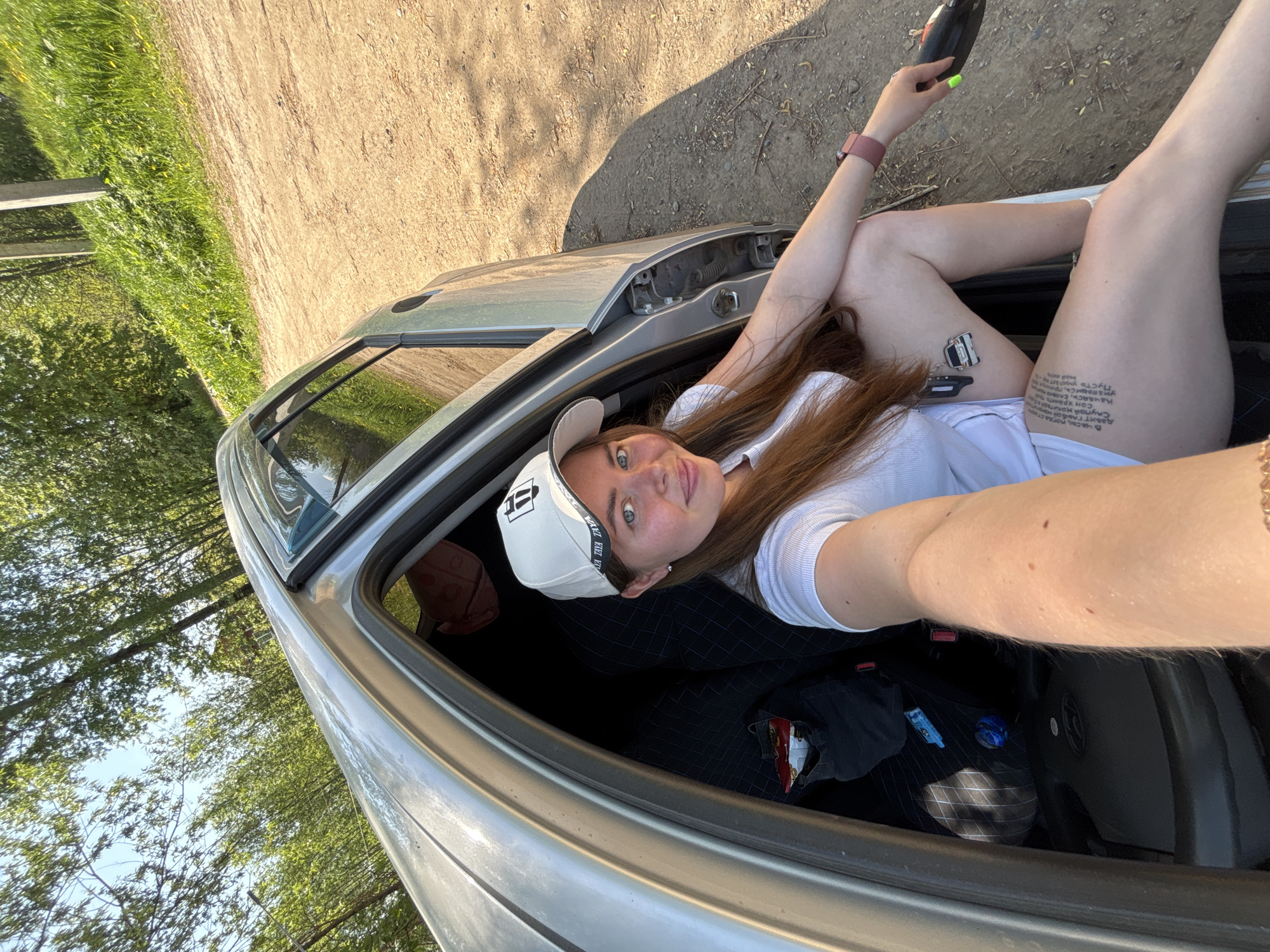 A woman with brown hair and blue eyes is posing inside the open trunk of a silver car. She is wearing a white T-shirt, a white baseball cap, and has visible tattoos on her right thigh. She is holding a black object in her right hand, and is wearing a pink smartwatch on her left wrist. The trunk interior is visible, containing clothes and other items. Outside the car, there is vegetation and a dirt road. The location is possibly Malyi Bukor, Russia.