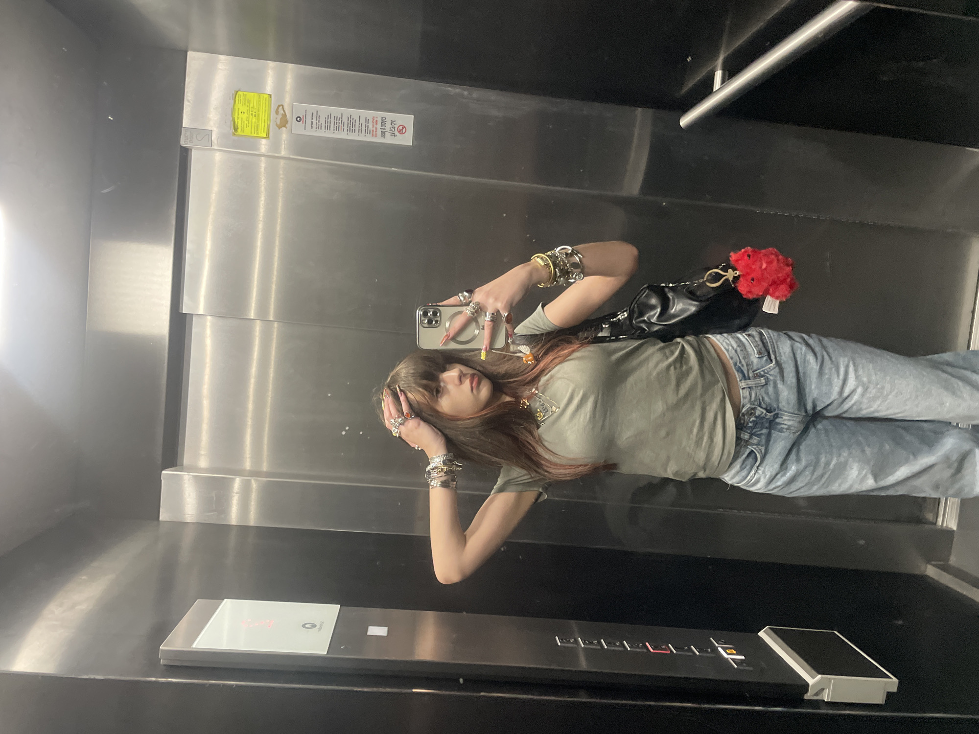 A young woman is captured in a mirror selfie inside a stainless steel elevator. She stands facing the camera, which she holds in her right hand, reflected in the polished elevator door. Her long, dark hair with reddish streaks falls over her shoulders, and she has bangs across her forehead. She is wearing a light olive green t-shirt, light blue washed-out jeans, and a black shoulder bag with a silver chain strap. A fluffy red pom-pom keychain is attached to her bag.
Her hands and wrists are adorned with numerous rings and bracelets, some silver-toned, others gold. Her fingernails are long, with yellow polish visible on some. The smartphone she uses to take the photo is light-colored and features a MagSafe ring on its back.
The elevator interior has reflective stainless steel walls and doors. On the right side of the elevator, a control panel with illuminated red buttons and a digital display showing "G" (likely for Ground floor) and another showing "2" are visible. Above the control panel, on the elevator door, are various informational stickers. A prominent yellow sticker displays a "NO SMOKING" symbol and text written in Hebrew. Another white sticker next to it also contains Hebrew text, likely instructions or warnings. A small metallic plaque with the letter "S" is also affixed to the door. A metallic handrail runs along the back wall of the elevator. The floor is dark and reflective. The setting suggests an indoor urban environment, specifically in an elevator in Raanana, Israel.