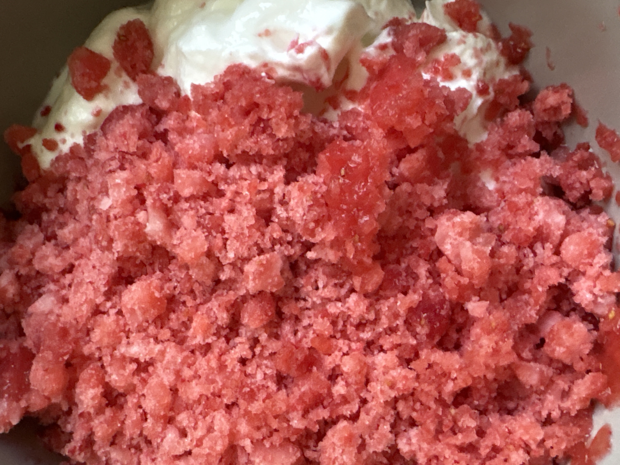 The image presents a close-up, overhead view of a food item composed of two primary components. The majority of the frame is filled with a coarse, granular substance, predominantly pink and reddish-pink in color. This material exhibits an uneven, somewhat icy texture, consistent with crushed fruit, granita, or a similar frozen dessert. Scattered throughout are smaller, lighter crystalline particles. To the right, a significant portion of smooth, opaque white material, resembling whipped cream, ice cream, or yogurt, is visible. Small fragments of the pink granular substance are interspersed within and resting on the surface of the white component. The background perimeter, visible mainly in the upper right, is a dark, indistinct surface, likely the interior of a bowl or serving dish. No human presence, specific actions, or environmental details are discernible that would allow for the determination of a geographical location, including the city of Saint Petersburg, Russia.