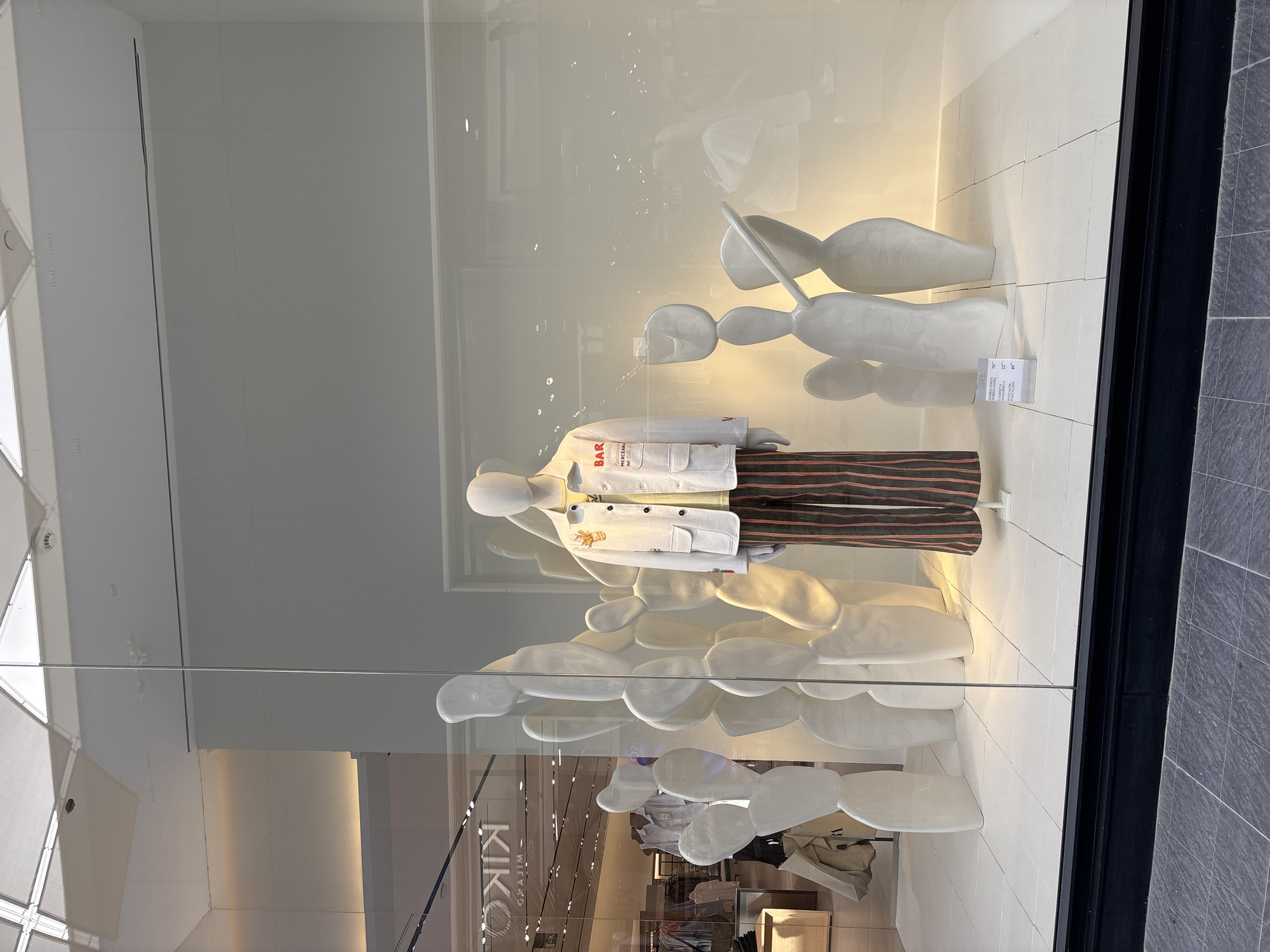 A mannequin is displayed in a shop window in Orihuela, Spain. The mannequin is wearing a white jacket over a yellow shirt and striped pants. Behind the mannequin are several white, abstract sculptures resembling cacti. The shop window displays clothing and accessories for sale. The background of the shop appears to be a retail store with racks of clothing.