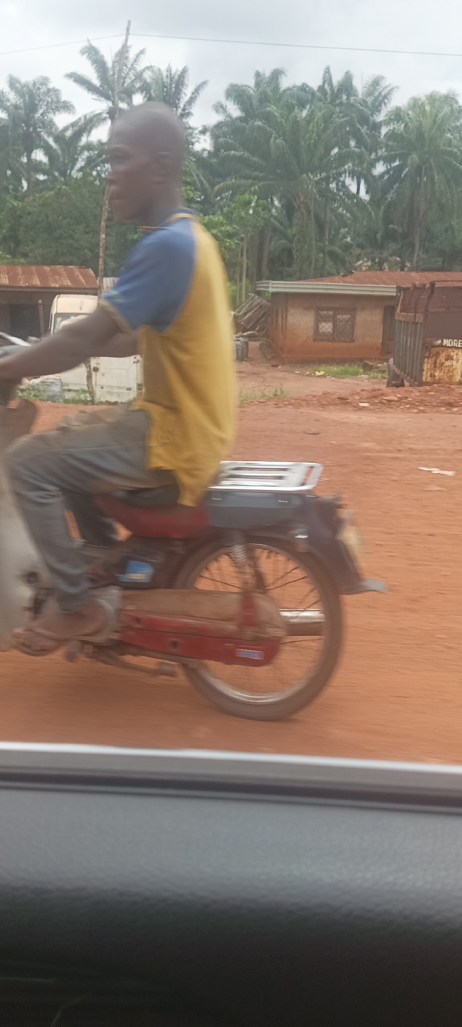 A man is riding a motorcycle on a dirt road in Imilike, Nigeria. The road is red dirt. The man is Black, bald, and wearing a yellow and blue shirt with blue pants. In the background are trees and a few buildings. The photo appears to have been taken from inside a vehicle.