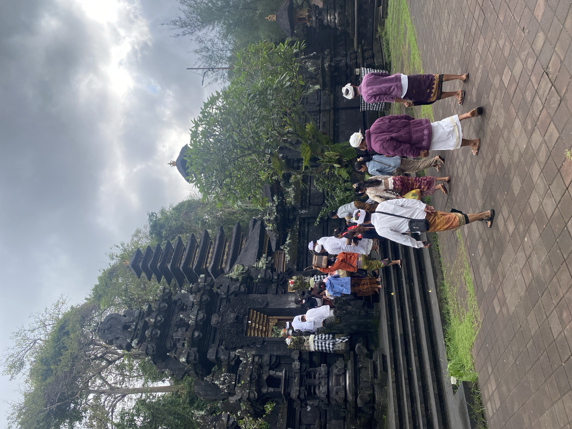A group of people, dressed in traditional Balinese attire, are walking towards and up the steps of a stone temple in Dawan, Indonesia. The temple features intricate carvings and tiered roofs, with a larger, multi-tiered structure to the left and a domed building in the background. Some individuals are wearing white shirts and sarongs, while others are in purple jackets and sarongs. The ground is paved with reddish-brown tiles, and patches of green grass are visible. The sky is overcast with gray clouds.
