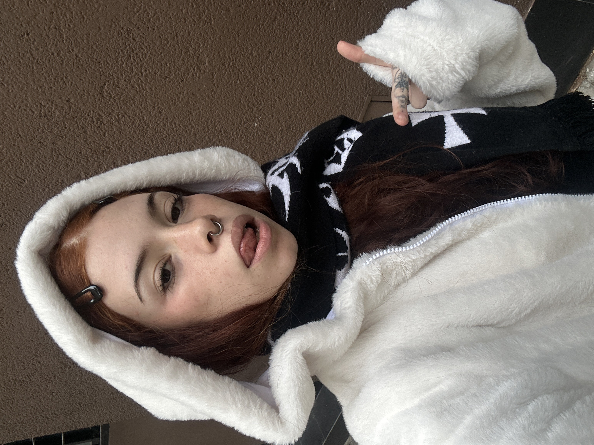 A young woman with red hair, freckles, and a nose ring is posing for a selfie. She is wearing a white, furry hooded jacket over a black and white scarf. Her left hand is raised with a tattoo visible on her finger. She is sticking out her tongue. Her hair is held back with a black clip. The background is a brown wall.
There is not enough information to determine that the photo was taken in Buenos Aires, Argentina.