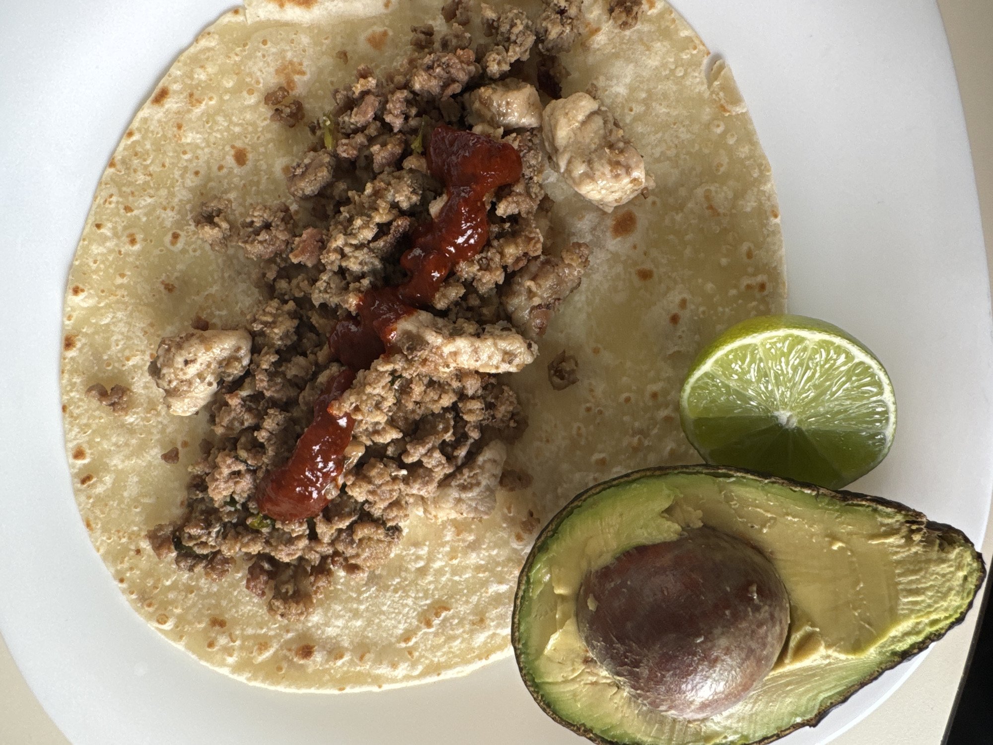 On a white plate, there is a flour tortilla topped with ground meat, cubes of light-colored meat, and a stream of red sauce. Next to the tortilla are a lime wedge and a halved avocado with the pit still in place. The location is not discernible from the image.