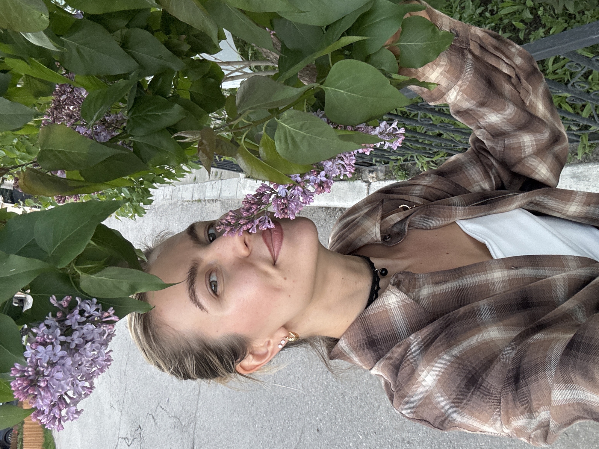 A young woman is photographed at eye-level with her head tilted to the left, her expression playful as she sticks her tongue out slightly. She is outdoors in what appears to be an urban environment, standing next to a flowering lilac bush. The bush's green leaves and clusters of light purple flowers frame the left side of her face.
The woman wears a brown and white plaid flannel shirt over a white top. She has fair skin, blonde hair pulled back, and a dark choker necklace. A gold hoop earring is visible in her left ear.
The background includes a section of gray sidewalk and a black metal fence partially obscured by foliage. The location is specified as the city of Unknown, Unknown.