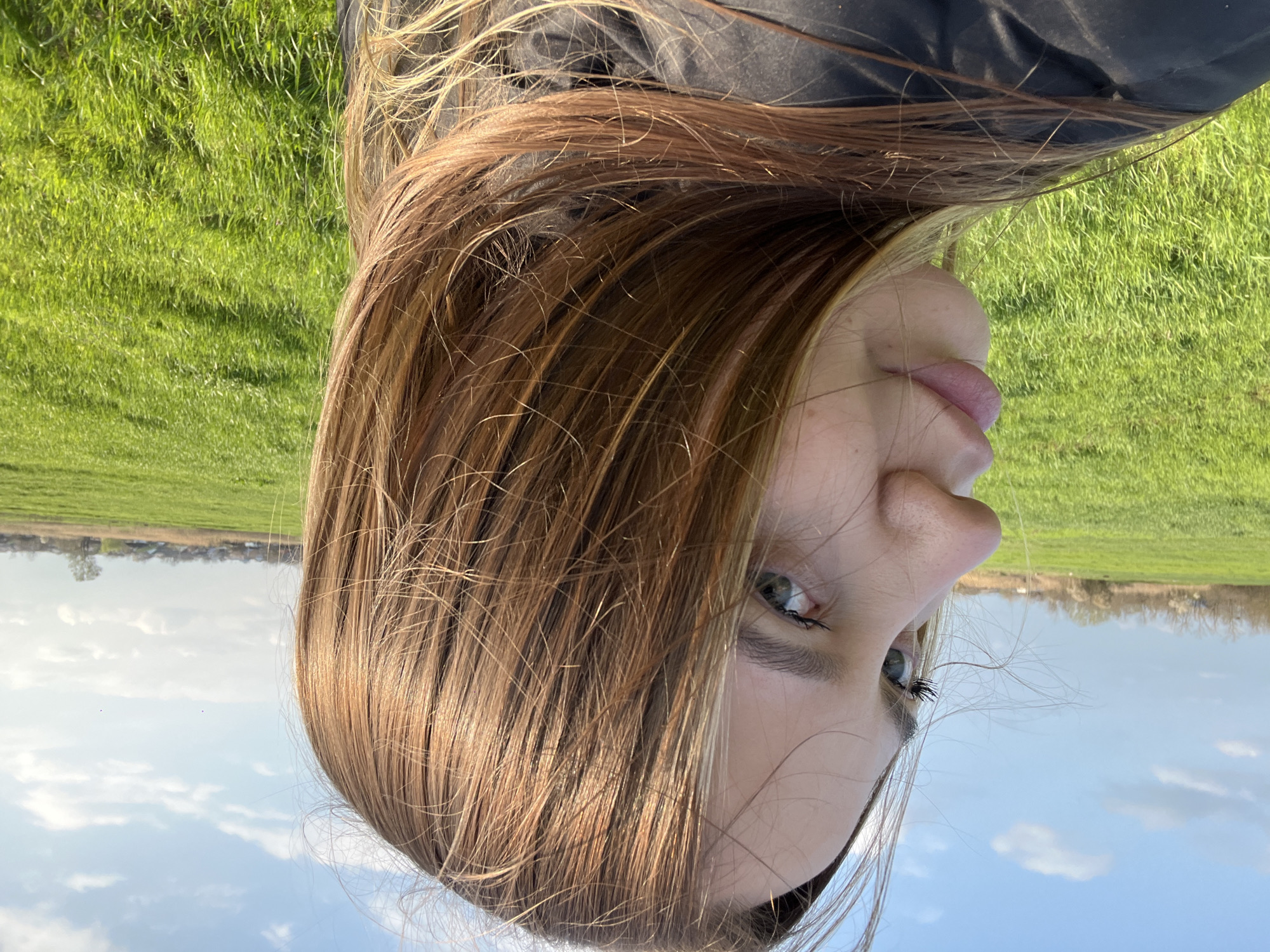 The image depicts a person lying in a grassy field, likely outdoors, with their head tilted towards the sky. The person has light brown hair and fair skin. They are wearing a dark-colored garment. The background features green grass, a blue sky with clouds, and distant buildings or trees along the horizon. The location is not discernible as Novopokrovskaya, Russia, solely from the provided image content.