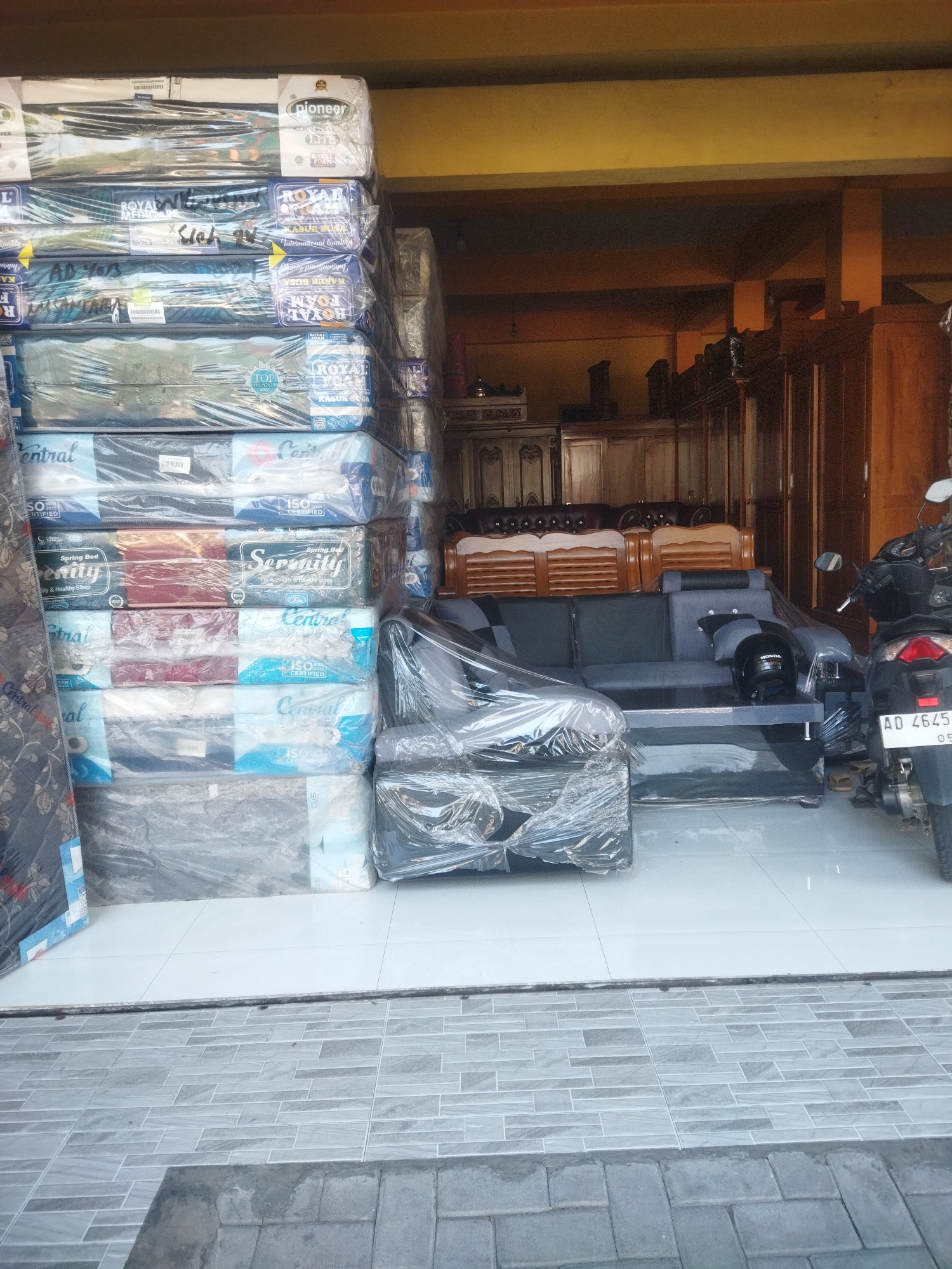 This is a photograph of a storefront in Sukoharjo, Indonesia, exhibiting a variety of mattresses and a sofa. In the foreground, stacked mattresses are visible, many wrapped in plastic. Brands such as "Pioneer," "Royal Foam," and "Central" are discernible on their packaging. Behind the mattresses, a grey sofa is also wrapped in plastic, with a black helmet placed on it. A motorcycle is parked to the right of the sofa. The background reveals wooden furniture, including cabinets and what appear to be chairs. The floor is tiled with light-colored tiles in the interior and grey brick pavers outside. The overall scene depicts a furniture and mattress retail establishment.
