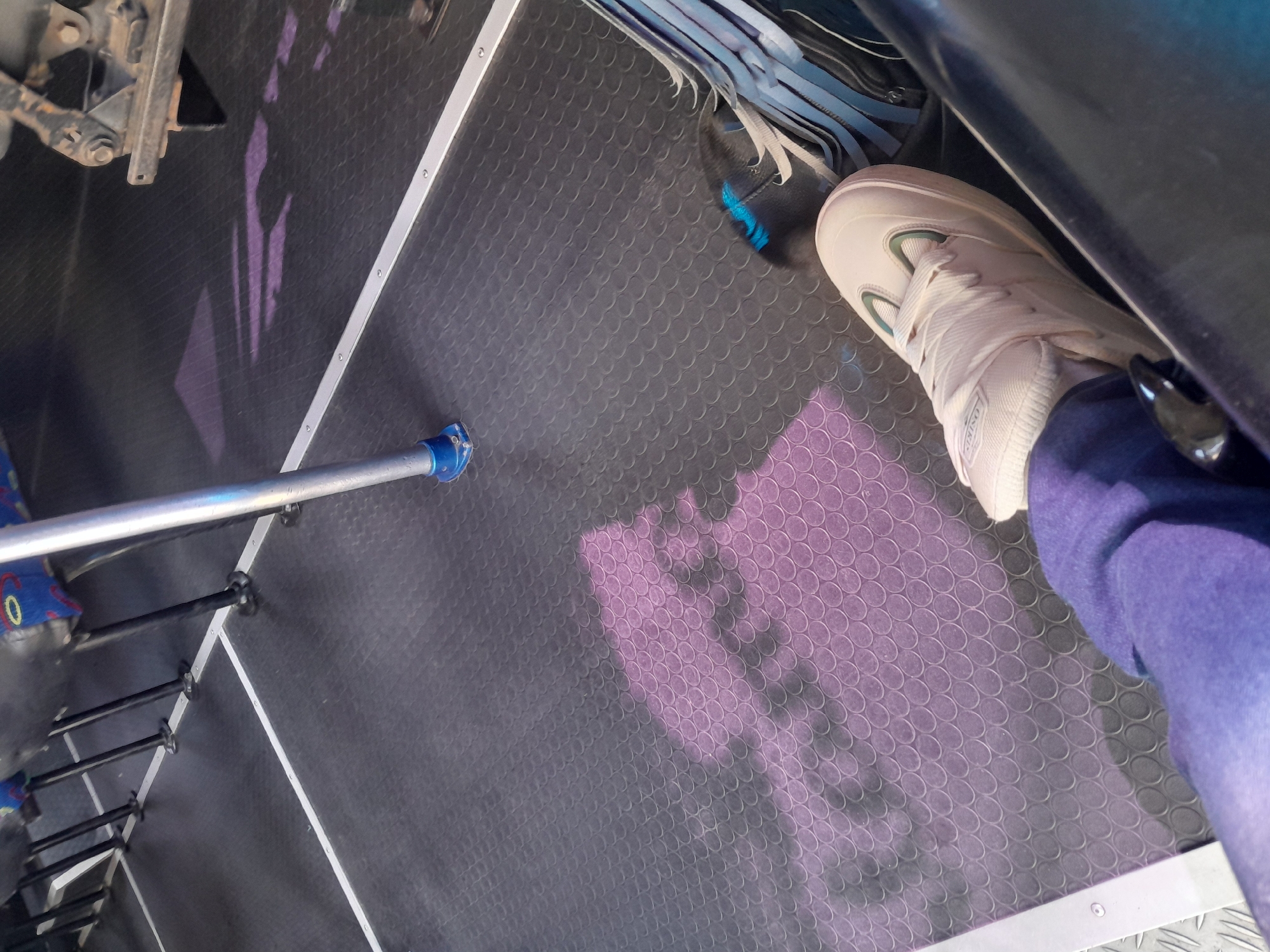 The image shows a close-up view of a person's feet inside what appears to be a bus or public transport vehicle. The floor of the vehicle is covered in a dark, textured rubber mat with a circular pattern. A metallic pole with a blue base is visible on the left side of the image. The person is wearing a light-colored sneaker and purple pants. A shadow with a patterned texture is cast on the floor, suggesting light is coming from above and to the side. In the background, the legs of several seats are visible, indicating a seating arrangement typical of a bus. The image does not provide any explicit details to pinpoint its location in Porlamar, Venezuela, or any specific city. The scene is a common interior view of public transportation.
