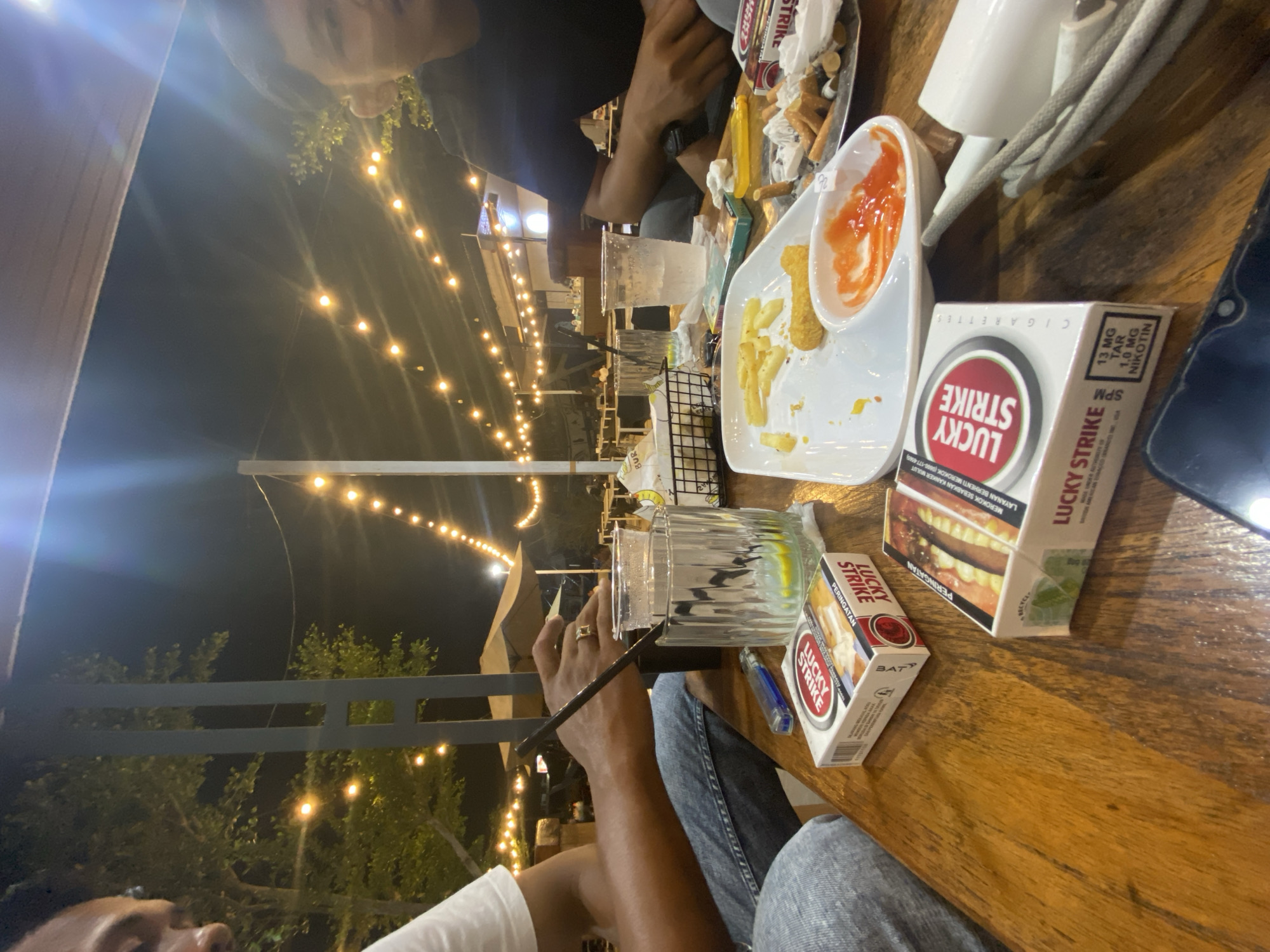 The image depicts a scene at night in Idi Rayeuk, Indonesia. There is an outdoor seating area illuminated by string lights. Two people are present at a wooden table. One person is wearing a black shirt and appears to be seated opposite another person wearing a white shirt. Food is present on the table, including a plate of fries and a dish with sauce. Two packs of Lucky Strike cigarettes are also on the table, along with a glass of liquid and a cell phone. A metal railing is visible in the background.