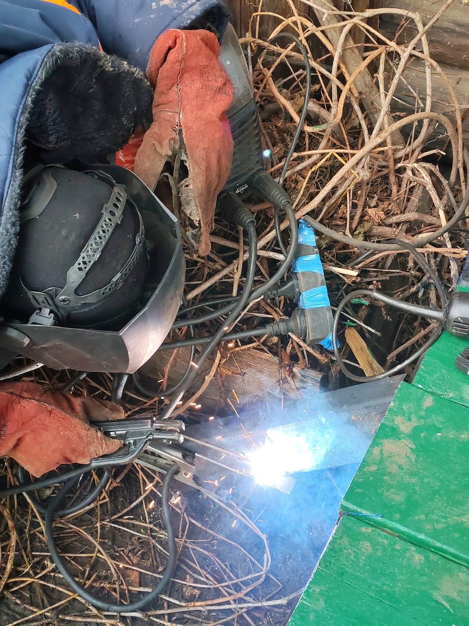 The image shows a welding operation in an outdoor setting. A welding helmet, gloves, and a jacket are present. Extension cords plugged into a power strip are also visible, and the welding equipment is actively being used, as evidenced by the bright light and sparks. The surrounding environment includes dry branches and wooden structures, possibly part of a building or fence. Without further information, a specific location within the city of Chaikovsky, Russia, cannot be determined.