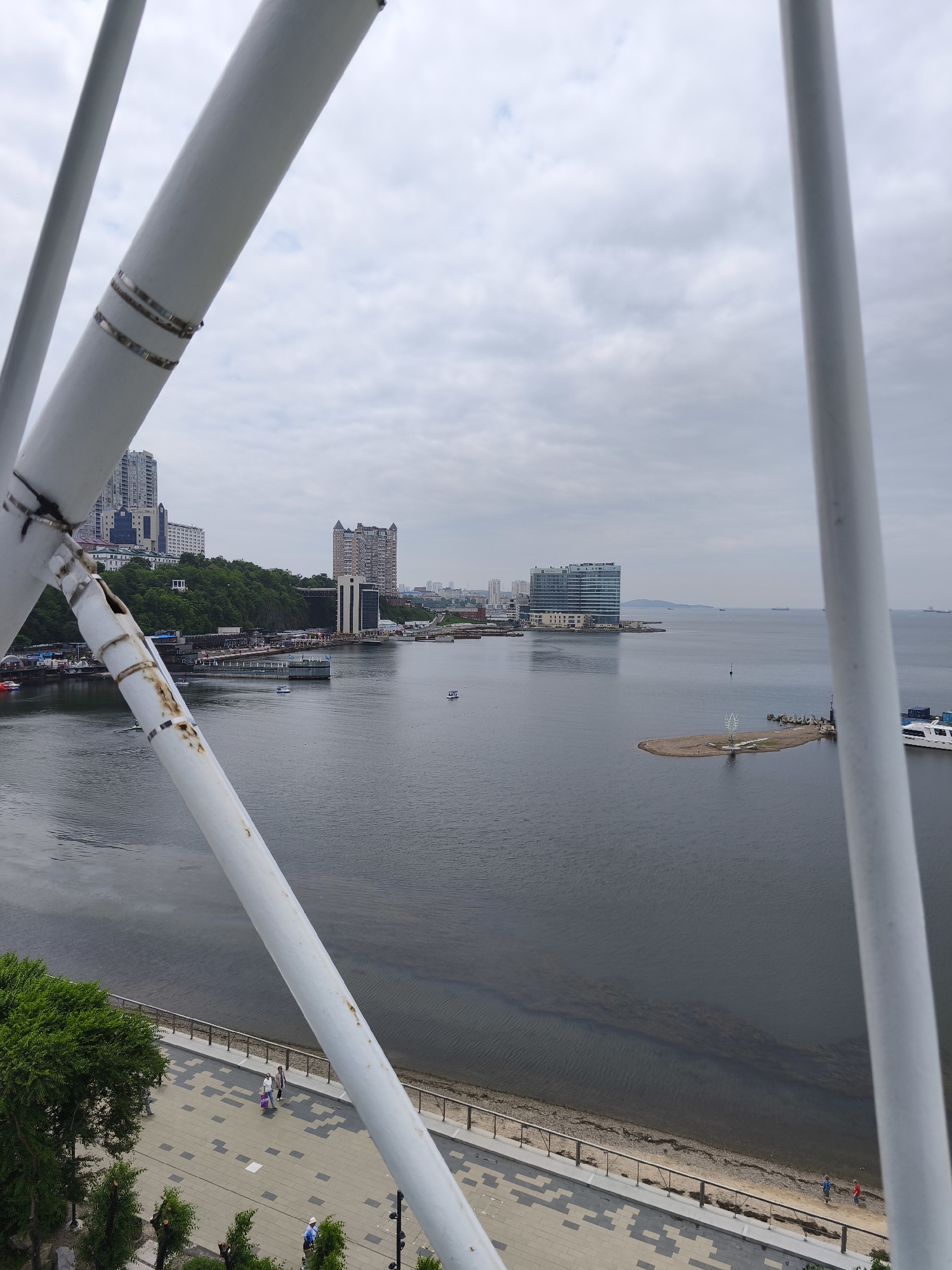 An elevated, medium shot captures a panoramic view of the coastline of Vladivostok, Russia, on an overcast day. The foreground is dominated by a portion of a white, tubular structure, possibly part of a Ferris wheel or observation deck, with visible metal bands and signs of rust. This structure frames the scene, creating a triangular opening through which the city and bay are viewed.
Beyond the foreground structure, the bay stretches out, with its dark water reflecting the muted sky. A paved waterfront promenade lines the edge of the bay, with a few people walking. Trees and some landscaping are visible along the promenade. Further out in the bay, various vessels, including boats and cargo ships, are present. A small, sandy island with a white structure is situated in the middle of the water.
The background features the Vladivostok cityscape, characterized by a cluster of modern and older buildings situated on a green, hilly landscape. A prominent modern building with a glass facade stands out on the left side of the frame, overlooking the bay. The sky is predominantly filled with gray clouds, suggesting a generally cool and possibly damp atmosphere. The overall scene depicts a typical day in a coastal city with urban development and natural elements.