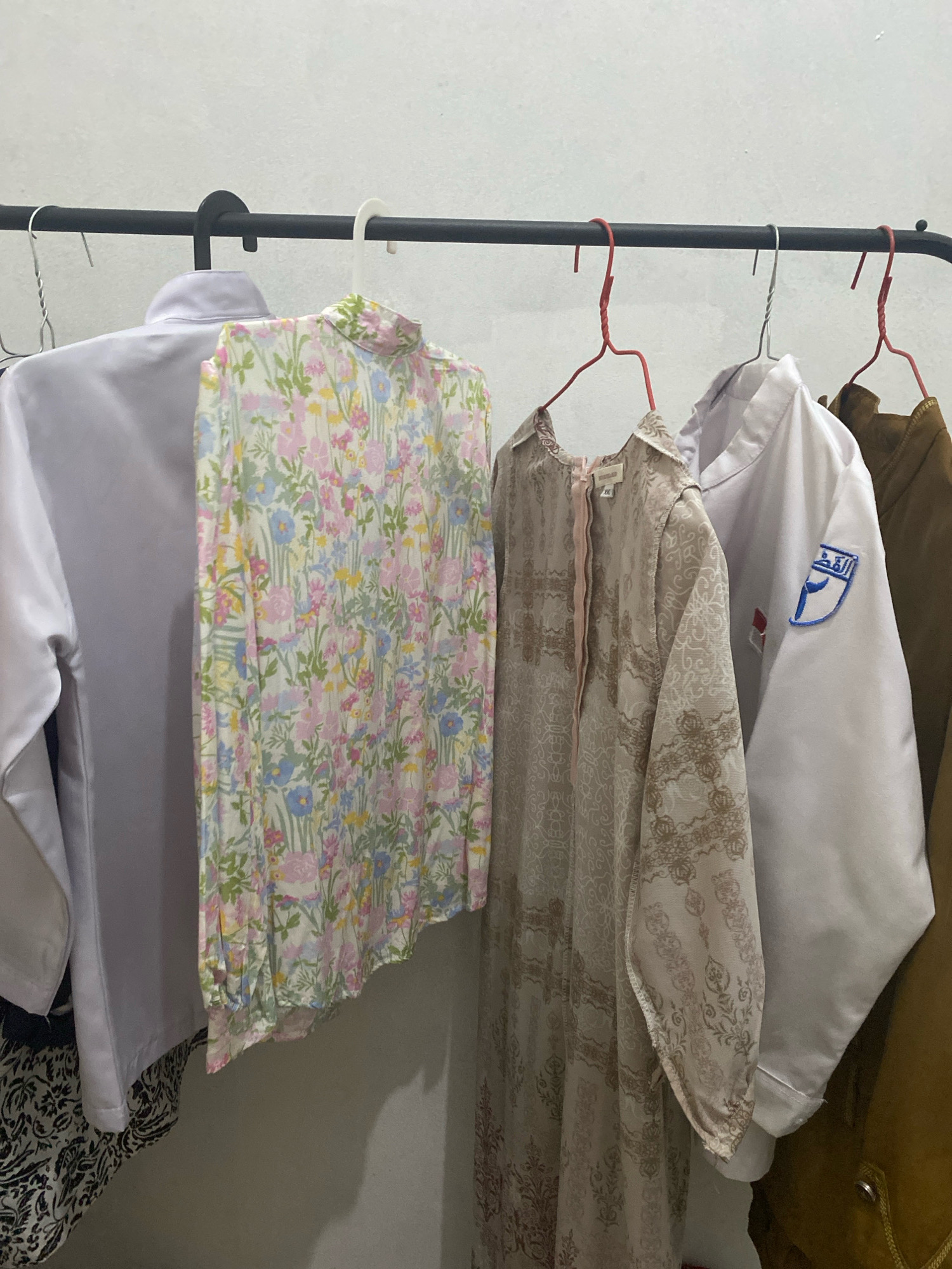 An indoor shot shows five garments hanging on a black metal clothing rack against a plain, white wall. From left to right, the garments are a white lab coat or uniform, a light-colored, long-sleeved blouse with a floral pattern in pink, blue, green, and yellow, a light brown, long-sleeved dress with an intricate pattern, another white lab coat or uniform with a blue and white logo on the left sleeve, and a brown jacket. The clothing rack is positioned in what appears to be a domestic setting, possibly a bedroom or closet, in Kota Dalam, Indonesia. The lighting is artificial, casting soft shadows on the wall. No people are visible in the image.