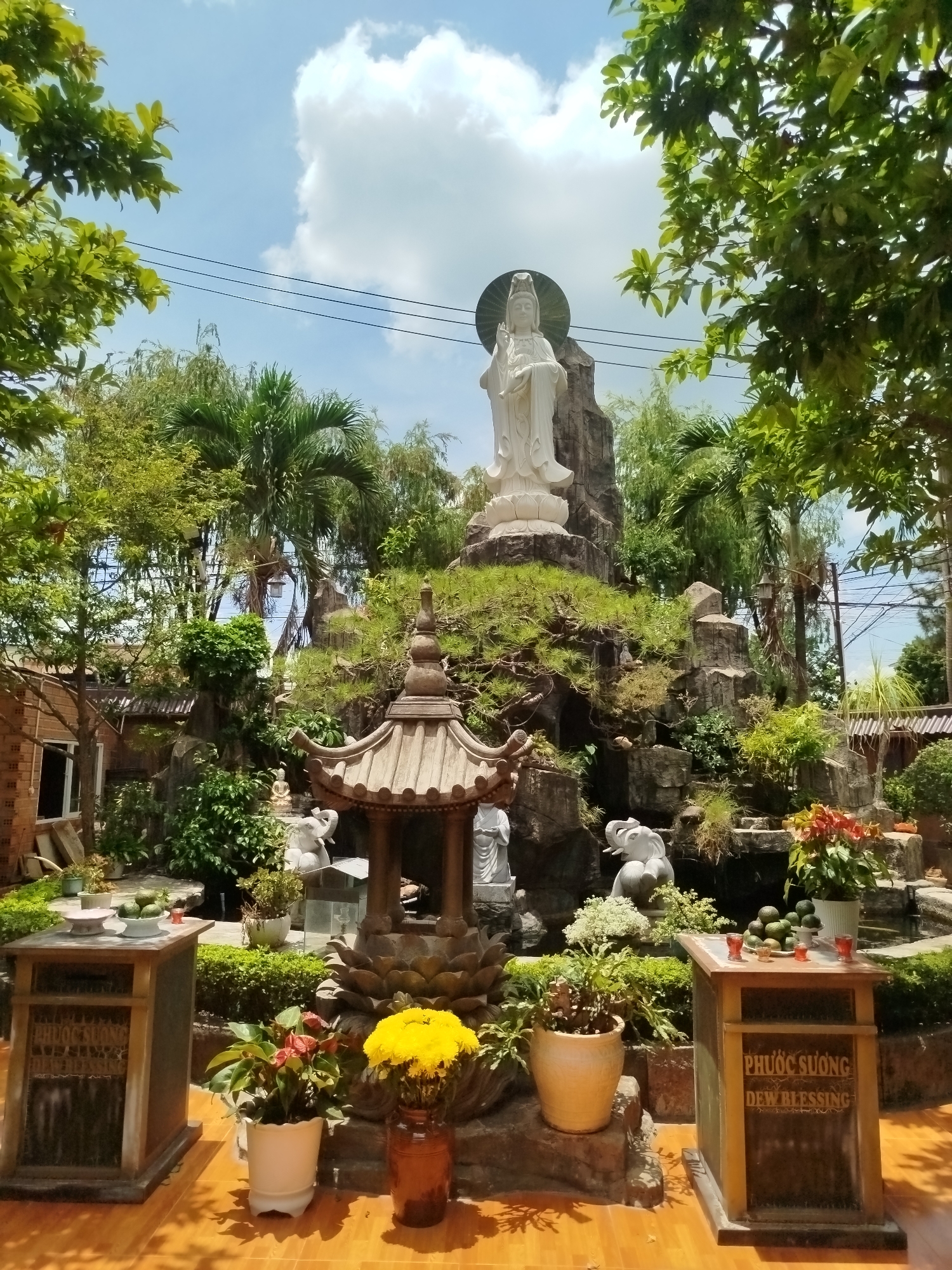 The image captures a religious scene in Dien Hong Ward, Vietnam. A prominent white statue of a deity stands atop a rock formation adorned with greenery. In the foreground, an ornate pavilion is surrounded by plants and flowers. Two wooden tables display offerings of fruit and candles. The surrounding environment includes trees and a blue sky with white clouds.