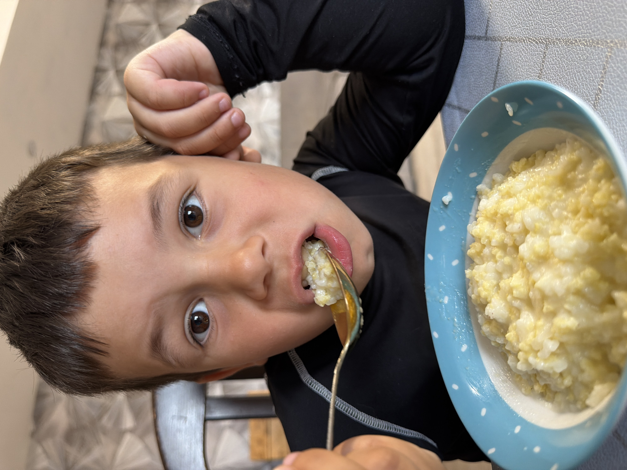 A young boy with dark hair and brown eyes is eating rice pudding from a blue and white polka dot bowl. He is holding a spoon to his mouth, which is filled with the pudding. The boy is wearing a black long-sleeved shirt. He is sitting at a table with a checkered tablecloth. The background is blurred, but it appears to be an indoor setting. The image was taken in the city of Chaikovsky, Russia.