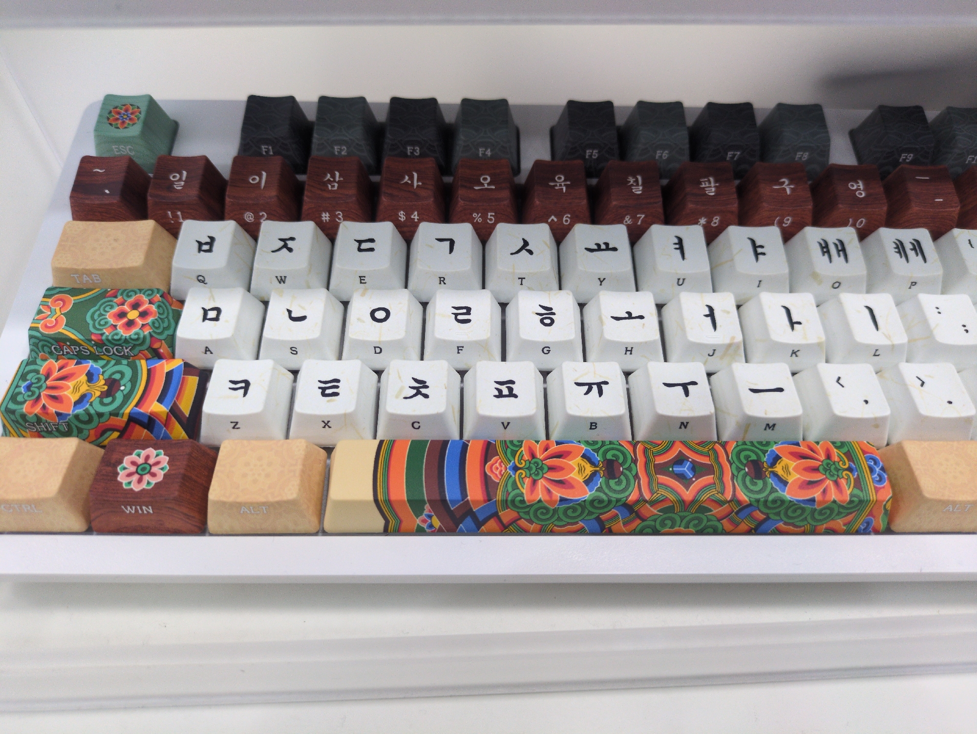 The image shows a keyboard with Korean characters on the keycaps. The top row of function keys (F1-F12) have a dark gray color with a wood grain texture. Underneath, there is a row of brown keycaps with Korean numbers printed on them (il, i, sam, sa, o, yuk, chil, pal, gu, yeong). The main alphabet keycaps are white with faint marble patterns and have both English and Korean letters. The Tab, Caps Lock, Shift, Ctrl, Win, and Alt keys feature wooden textures, with the Caps Lock and the spacebar featuring colorful traditional Korean patterns. The Caps Lock key has a flower design. The spacebar is decorated with a complex traditional Korean flower pattern. The overall setting is a bright, clean environment, suggesting an indoor location. The image provides no information on the specific location in Gyeongju-si or about people, actions, or interactions.