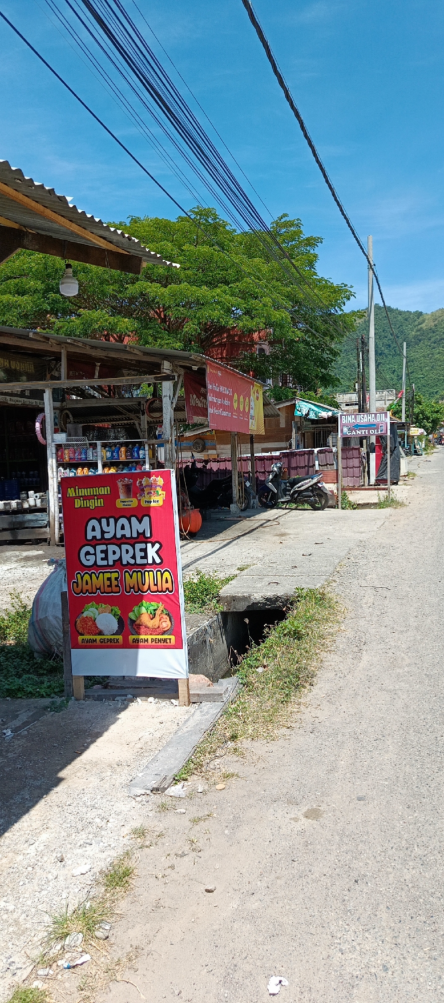 The image depicts a street scene in Rimajeunue, Indonesia, under a clear blue sky. On the left, a roadside stall displays various products, including beverages in bottles and cans, with a prominent red banner advertising "AYAM GEPREK JAMEE MULIA". This banner features images of "AYAM GEPREK" and "AYAM PENYET". Adjacent to the stall, a large white garbage bag is visible. Further down the street, a motorcycle is parked. The stall has a corrugated metal roof and wooden structures. On the right side of the street, a sign reads "BINA USAHA OIL GANTI OLI". The road surface is paved, with a concrete drainage channel running alongside it. Vegetation, including trees and grass, is present along the sides of the road and near the buildings. In the background, hills are visible. Power lines are strung across the top of the image.