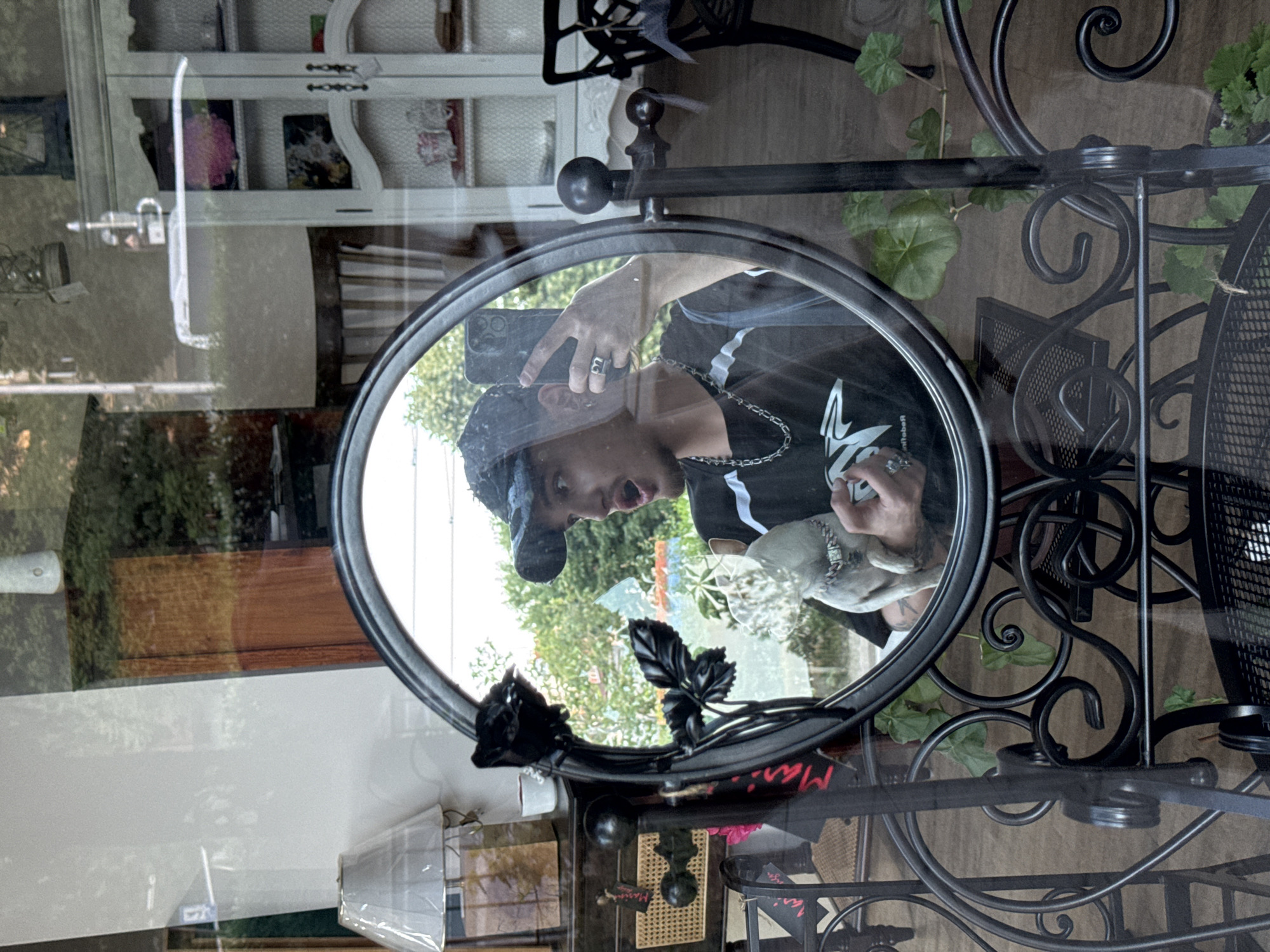 A medium shot shows the reflection of a young man taking a photo of himself in an oval mirror with a black metal frame. The mirror sits on a table with a black metal base with decorative swirls. In the reflection, the man wears a black baseball cap, a black t-shirt with white stripes on the shoulder, and a silver chain. He holds a mobile phone with a black case in his left hand and makes a surprised expression, his mouth open. He holds a small, decorative object resembling a dog or other animal in his right hand.
The reflection also shows a view through a window to a green outdoor area with trees and a building in the background. Behind the mirror, visible through the window and in the reflection, there is a white cabinet with shelves and various items on display. To the right of the mirror frame, green plants and a black metal chair are visible. The table is made of transparent glass.
There is no information on the location in Sofia, Bulgaria, within the image.
