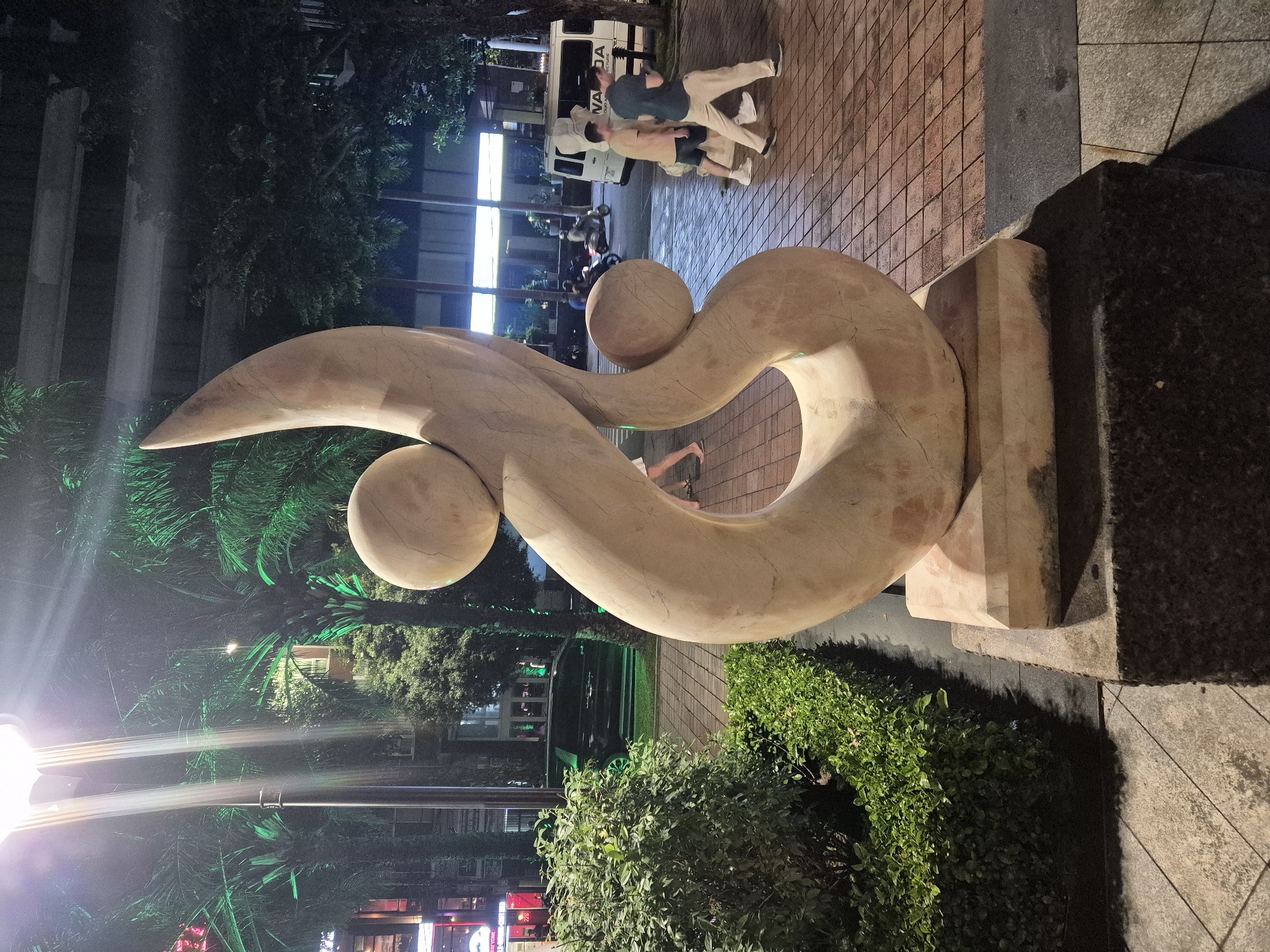 This is a low-angle, eye-level, outdoor, nighttime photograph taken in Da Nang, Vietnam. The central subject is a large, abstract, stone sculpture of a swirling, organic form with two spheres. The sculpture is positioned on a plaza with patterned red brick paving. To the left of the sculpture is a well-maintained green hedge and some palm trees illuminated by green lights. In the background, there are buildings with illuminated windows and signage. A black van is parked on the street behind the hedge, and several people are walking on the plaza. One person on the right is wearing a tan t-shirt and shorts, and another person next to them is wearing a dark t-shirt and light-colored pants. A white van is parked further down the street, and a motorcycle is visible in motion. A streetlamp with a bright light source is located to the left of the sculpture. The scene appears to be a pedestrian area in a commercial or entertainment district of the city.