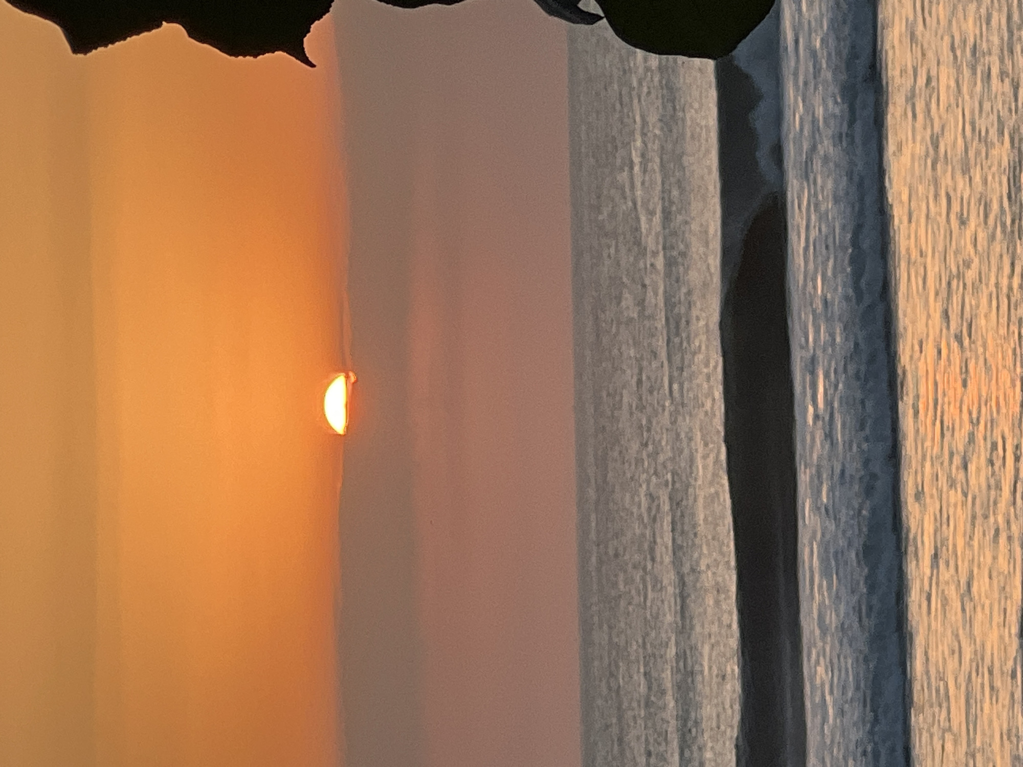 A telephoto view captures a sliver of the sun setting over the ocean, partially obscured by a band of hazy clouds. The sun, a glowing orb of bright orange and yellow, is situated near the horizon. The sky transitions from a vibrant orange at the top to a softer pink and peach hue as it nears the ocean. The water is calm with gentle waves breaking closer to shore, reflecting the warm colors of the sunset. The scene is framed on the right by the dark silhouette of a person or large object, possibly a statue or a tree, adding a dramatic contrast to the vibrant sky and sea. The location is identified as Kuta, Indonesia.