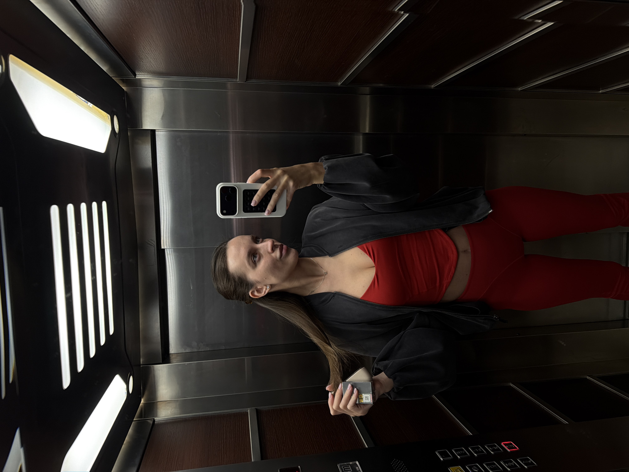 A young adult woman is seen taking a mirror selfie inside an elevator. The image is presented horizontally, though the scene depicts her standing upright within the elevator car. She has long, straight brown hair and a neutral expression, gazing towards her phone screen.
She is dressed in a vibrant red athletic crop top and matching red leggings, layered with a loose-fitting dark grey or black jacket. Her fingernails are painted light pink. In her right hand (from her perspective), she holds a white smartphone with a prominent black camera module, seemingly capturing her reflection. Her left hand holds a small, light-colored rectangular box, possibly an electronic device, featuring a visible QR code and some indistinct text, possibly "FLUX."
The elevator interior features shiny, brushed metal walls and a ceiling with dark brown wood-paneled sections. Bright, vertical strip lights illuminate the space intensely from the left side, casting reflections on the metallic surfaces. The floor appears to be a dark wood or laminate. At the bottom of the frame, the elevator control panel is visible, equipped with numerous buttons for different floors, including an illuminated red "1" button. Small text that appears to be "LUX" is also visible on the panel. The setting is an elevator, consistent with typical modern buildings, and is contextually located in Budva, Montenegro.
