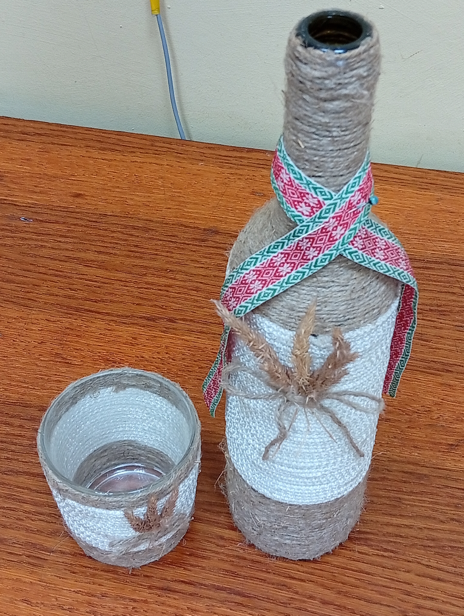 The image shows a decorative bottle and a glass, both covered in twine and a white fabric, sitting on a wooden surface. The bottle is tall and slender, with twine wrapped around it from the base to the neck. A festive ribbon with red, white, and green patterns is tied around the upper portion of the bottle, crossing in the front. A small bunch of dried grass is attached to the white fabric covering the lower half of the bottle. To the left of the bottle is a small glass, also wrapped in twine and white fabric, with a similar dried grass embellishment at its base. The background is a textured, light-colored wall, with a hint of a yellow and blue wire visible at the top left. The wooden surface appears to be a table. The scene is indoors, likely in a domestic setting within Крыўлянскі сельскі Савет, Belarus. There are no people or animals present in the image. The lighting is even, suggesting an indoor light source.
