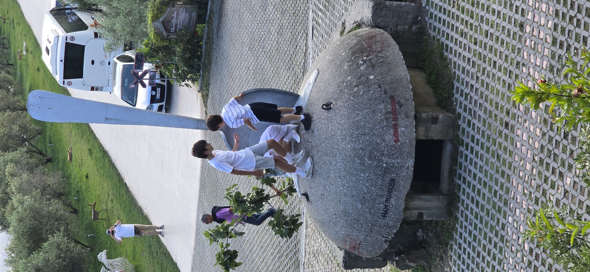 The image depicts a scene near the "Duhra Dashni" sign in Blinisht, Albania. A concrete structure resembling a bunker or dome is prominently featured. Two boys are seen on top of the structure. A man is also near the edge of the building, with another man behind the boys. Additionally, a car and a white bus are parked alongside a path. The surrounding environment includes a grassy area with trees and a paved walkway.