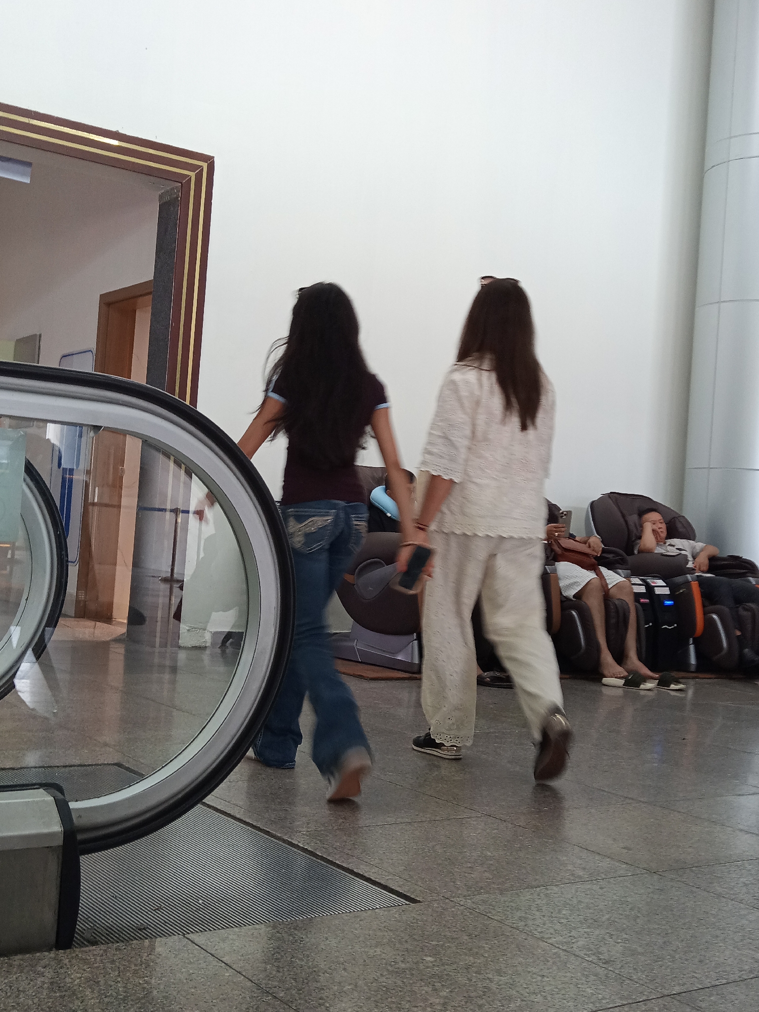 The image captures an interior scene, likely an airport lounge or a waiting area, given the presence of massage chairs. Two women are walking away from the camera, one in a dark top and blue jeans, and the other in a white lace top and white pants. In the background, a man is seated in a massage chair, appearing to be resting or asleep. Another person is partially visible in a massage chair to his left. An escalator's handrail and glass casing are prominent in the foreground, indicating a transportation hub. The environment features a polished stone floor and light-colored walls, suggesting a modern and clean facility. While the specific location in Đà Nẵng, Vietnam is not explicitly shown, such amenities are commonly found in its international airport or major hotels catering to travelers.