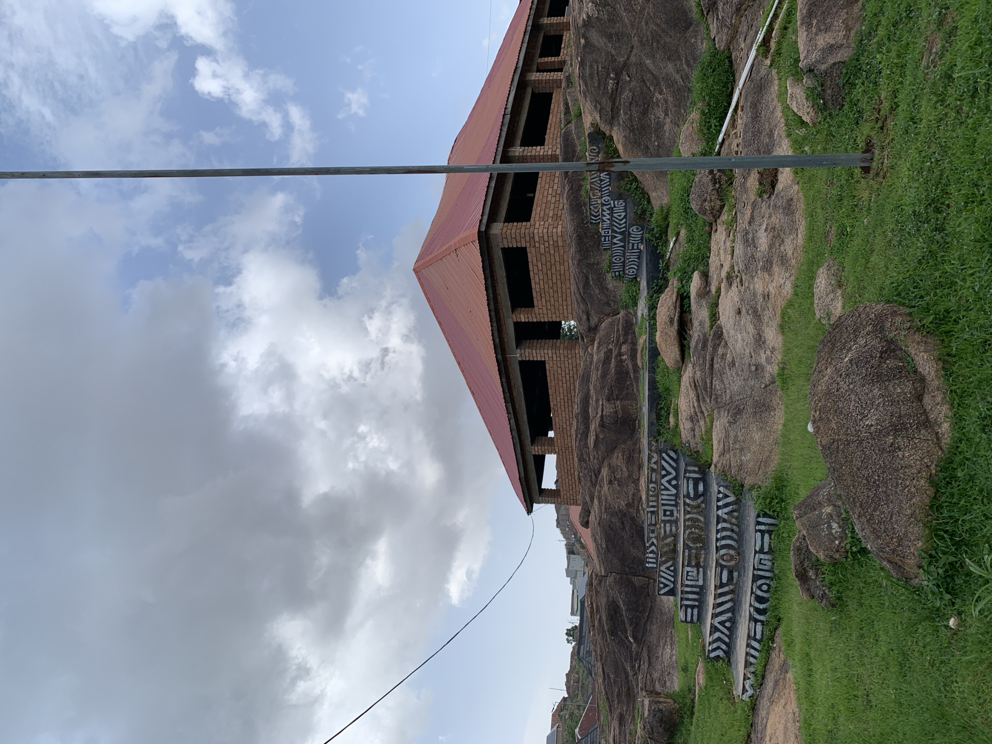 In Jos, Nigeria, a brick building with a red corrugated metal roof stands on a rocky outcrop. The building is open-sided, with visible brick walls and no doors or windows. A set of concrete stairs, decorated with black and white geometric patterns, ascends towards the building. The stairs are situated on a rocky landscape covered in patches of green grass. In the foreground, large boulders are scattered across the grassy terrain. A tall, thin metal pole stands vertically to the right of the stairs, with a thin black wire stretching diagonally across the sky from the left side of the frame towards the building. The sky is partly cloudy, with a mix of gray and white clouds. In the background, other buildings and structures are visible, suggesting an urban or semi-urban environment.