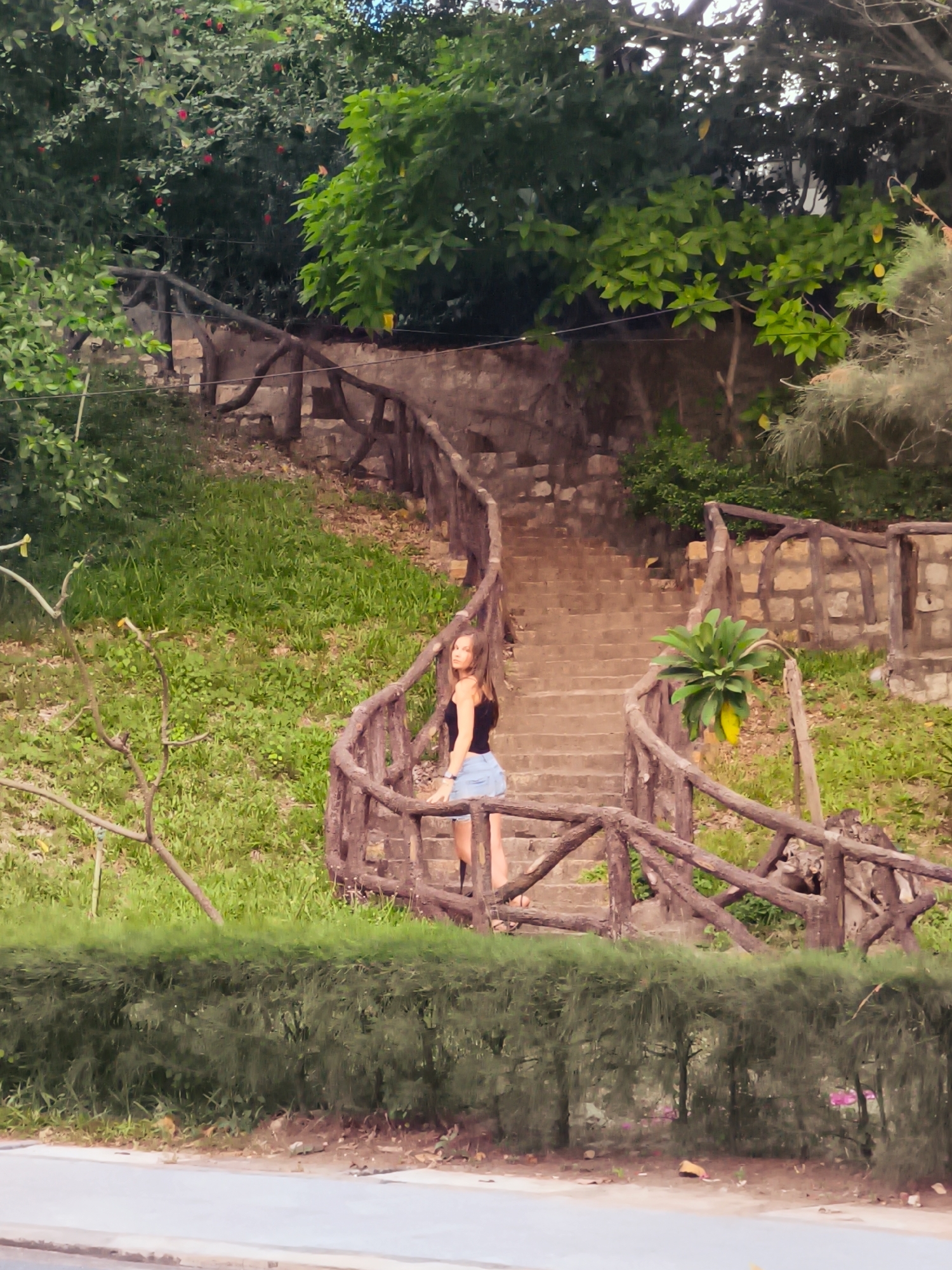 A young woman with blonde hair, wearing a black tank top and denim shorts, stands on a stone staircase with a rustic wooden railing. She is positioned on the lower half of the staircase, looking towards the camera. The staircase is surrounded by lush green grass and trees, with a stone wall visible behind the railing. The scene appears to be an outdoor park or garden area. The location is identified as Bac Nha Trang Ward, Vietnam.