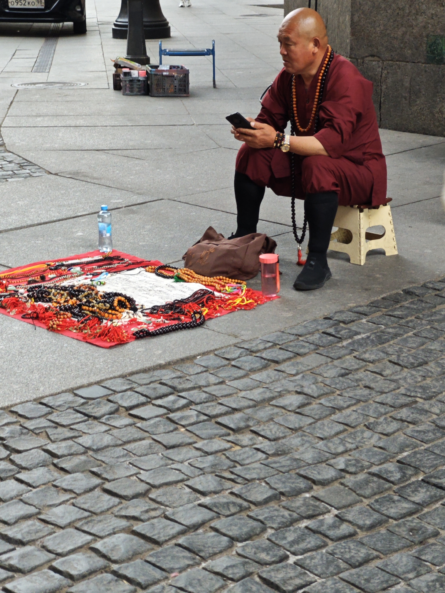 A bald man, dressed in a maroon robe and black leggings, sits on a small, cream-colored stool on a cobblestone sidewalk. He is looking at a black smartphone in his hand. Around his neck, he wears multiple strands of beaded necklaces. To his left, a red cloth is spread on the ground, displaying a variety of beaded bracelets and necklaces. A bottle of water and a pink plastic container with a red lid are placed near the display. To the man's right, a brown bag rests on the ground. In the background, a blue metal bench is visible, along with several plastic crates. A black car is parked on the left side of the image. The scene takes place in Saint Petersburg, Russia, on a city sidewalk with cobblestone paving.