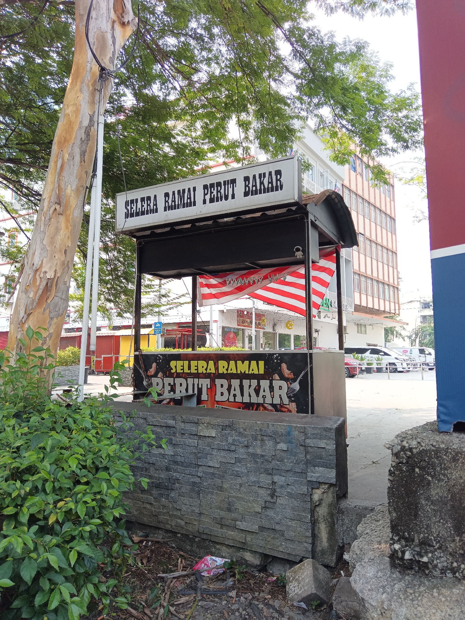The image captures a food stall located in Kuala Lumpur, Malaysia, specifically outside a building complex. The stall, which appears to be a permanent structure, features a prominent sign in Malay reading "SELERA RAMAI PERUT BAKAR" (meaning "Many Appetites Grilled Stomach"). This sign is displayed both at the top of the stall and on its front panel, which is adorned with graphics of grilled food.
The stall has a dark canopy and is decorated with a Malaysian flag. In front of the stall, a stone-clad wall with a textured finish forms a barrier. Behind the stall, a modern, multi-story building with many windows is visible, suggesting an urban setting. The surrounding environment includes trees with lush green foliage, a paved walkway, and some parked vehicles in the background. The ground area near the stall is somewhat cluttered with debris, including some discarded plastic packaging. The overall scene depicts a commercial streetscape in Kuala Lumpur on a bright, possibly sunny day.