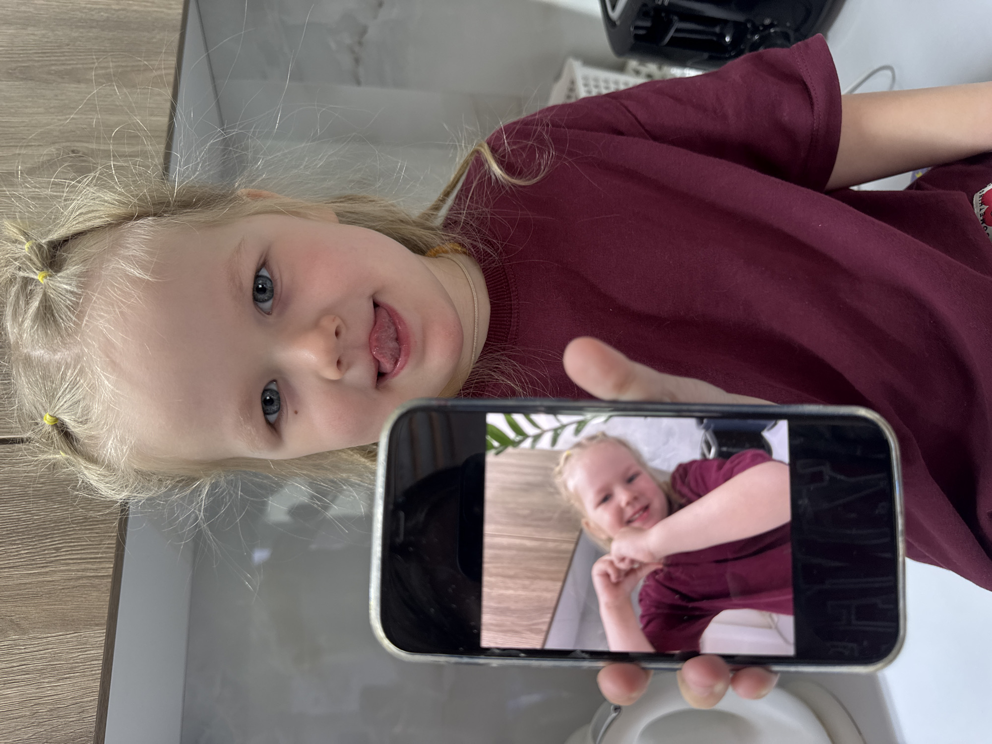 A young blonde girl with her hair tied in pigtails is holding up a smartphone that displays a photo of herself smiling and posing with crossed arms. She is wearing a maroon t-shirt and sticking her tongue out. The scene appears to be inside a bathroom, with a toilet and other bathroom fixtures visible. The location is described as Gornyi (Dvurechenskoe S/P), Russia.