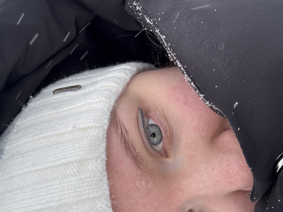 The image depicts a close-up, angled view of the upper left portion of a person's face, suggesting an outdoor winter environment in Nizhnekamsk, Russia.
**People:**
A single individual is partially visible. The subject's left eye is prominent, featuring a light blue iris with a visible pupil, and a thin line of dark eyeliner on the upper eyelid extending slightly past the outer corner. The eyebrow above is light brown and appears natural. The skin around the eye and on the visible part of the cheek and nose exhibits a somewhat rosy, reddened appearance, consistent with cold exposure. Fine facial hair is visible on the cheek and forehead.
**Subjects/Objects:**
The person wears a white, ribbed knitted beanie on their head, part of which is visible above the left eyebrow and temple. A small, dark, rectangular metal tag or embellishment is affixed to the right side of the beanie. The lower portion of the face, including the mouth and chin, is obscured by the dark, possibly black, collar or hood of a thick, padded winter jacket, which appears to be made of a synthetic, slightly textured fabric. The jacket material is visible on the right side of the frame, wrapping around the lower part of the face. A metallic clasp or zipper pull is partially visible at the bottom edge of the jacket, near the chin area.
**Actions/Interactions:**
The person appears to be facing slightly forward, with the jacket pulled up to cover a significant portion of the lower face, indicating an attempt to shield from cold. Scattered small, white flakes, consistent with snowflakes, are present on the beanie, the visible skin, and the fabric of the jacket.
**Environment:**
The background directly behind the person's head and neck, within what appears to be a hood or shadowed area, is dark and indistinct. The presence of snowflakes on the clothing and skin indicates active snowfall and cold weather conditions.