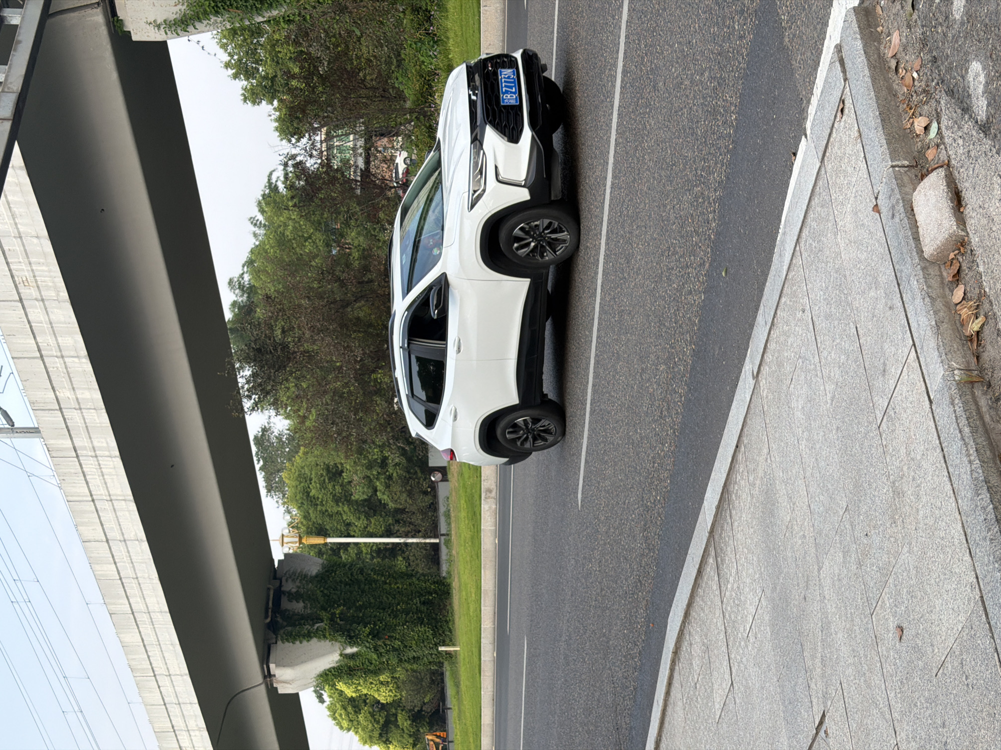 A white Chevrolet Trax is driving on a paved road in Jiangjin, China, on a cloudy day. The car is moving from left to right, with its front facing forward. The road is wet from recent rain, and there are painted white lines on the asphalt. To the left of the road is a sidewalk made of gray concrete tiles, bordered by a concrete curb. Beyond the sidewalk is a grassy area with a yellow lamppost. In the background, a large concrete overpass with railway tracks runs horizontally across the image. Lush green trees and vegetation grow beneath and around the overpass, with some trees showing signs of autumn colors. The sky is overcast and light.
