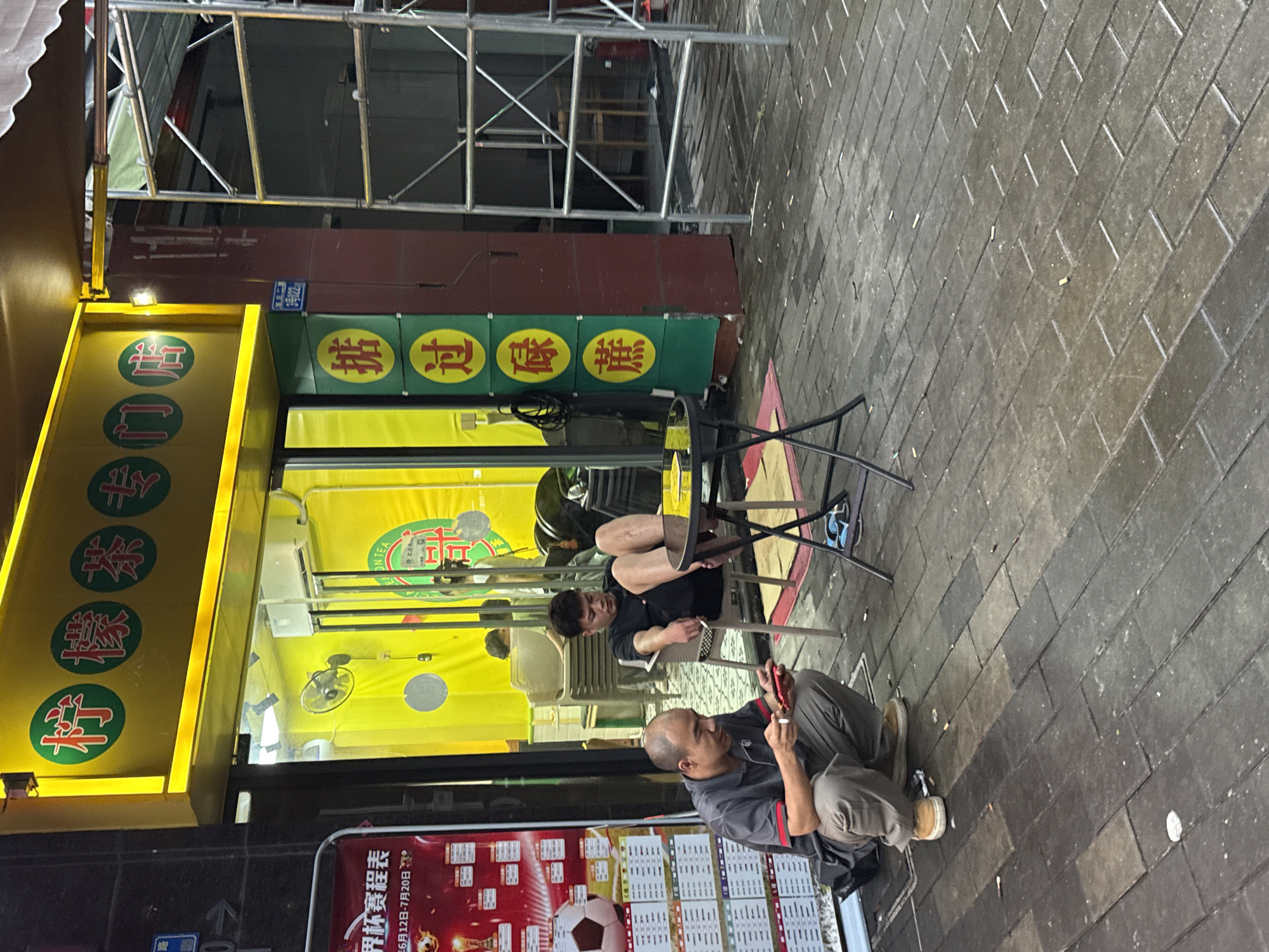 The image depicts a street scene in Huizhou, China, likely in the evening. In the foreground, a man is squatting on the pavement, looking at a red mobile device and holding a cigarette. He is wearing a grey polo shirt and grey trousers. Next to him, a young man sits in a chair, looking down at his phone. He is wearing a black t-shirt and black shorts. A small black table with a glass top stands between them. The pavement is made of dark interlocking bricks, many of which are wet and reflect the light. Scattered on the pavement are cigarette butts.
Behind the men, the entrance to a shop is visible. The shop has a bright yellow facade with a prominent sign featuring Chinese characters in red circles. A narrower green sign with red Chinese characters stacked vertically is also present. The interior of the shop appears to be brightly lit with yellow walls and a fan on the wall. Further back, scaffolding for construction or renovation is visible. The overall atmosphere suggests a busy urban environment, possibly a commercial street.