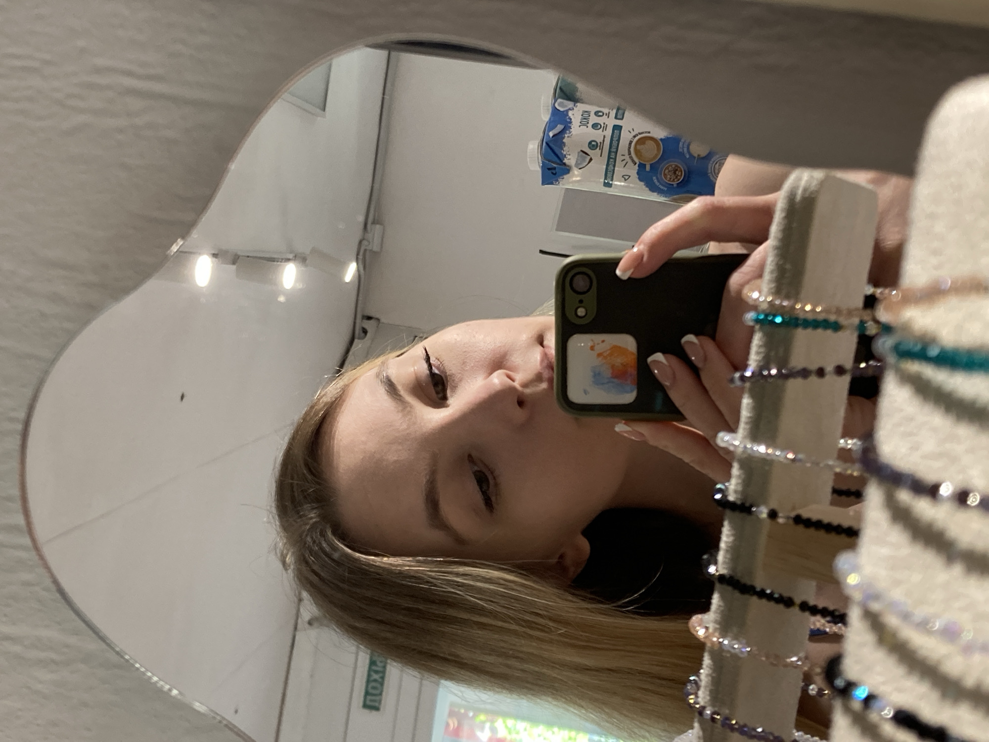 A mirror selfie photograph shows a woman reflected in a mirror, holding a cell phone with a dark green case. Her hair is blonde with visible dark roots. She has a light complexion and is wearing eyeliner. Her manicured nails have a white tip design. The reflection reveals part of a room with a white wall, a sign in what appears to be Russian, and a carton of what looks like coconut milk. In front of the woman are bracelets displayed on a light-colored stand. The location is inferred to be in Chaikovsky, Russia.