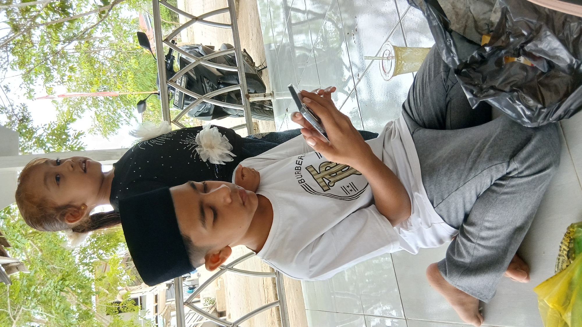 In Matangben, Indonesia, two young individuals are captured in a portrait. A young boy, wearing a black peci (a traditional cap) and a white t-shirt with a graphic design, sits on a tiled floor. He is engrossed in a mobile phone he holds in his hands. Beside him, a young girl, with her hair in a ponytail adorned with white flowers, stands and looks to the right. She is wearing a black top embellished with small sparkling details. Behind them, a motorcycle is visible, and a metal railing borders a raised platform. The environment appears to be an outdoor shaded area, possibly a veranda or a covered walkway, with trees and greenery in the background.