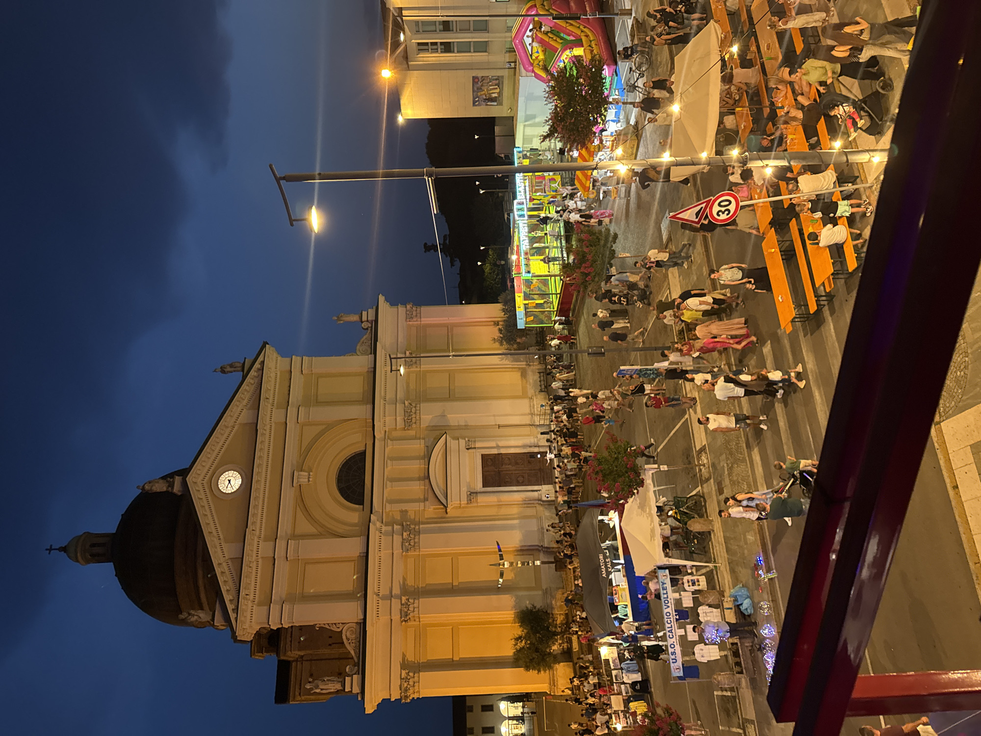 The image depicts an evening outdoor event in the city of Calcio, Italy, centered around the town's church. The church, a prominent yellow building with a large dome and a clock face, is illuminated. The scene is busy with people of all ages, many of whom are standing, walking, and sitting at tables.
In the foreground, a red railing suggests the viewpoint is from an elevated position, possibly a balcony or higher floor. The ground level features market stalls, one with "U.S.O. CALCIO VOLLEY" printed on it, selling merchandise. Further into the scene, there are inflatable attractions, including a bouncy castle, and a colorful trampoline-like structure labeled "TAPPETI ELASTICI". Orange tables and benches are set up, indicating a dining or gathering area, with many people seated and interacting.
Several streetlights and lights from the stalls illuminate the area, creating a lively evening atmosphere. A "30" speed limit sign is visible, suggesting the event is taking place in a public square or street that is temporarily closed or managed for the occasion. The sky is a deep blue, indicating it is dusk or early night. The overall impression is of a community festival or fair.