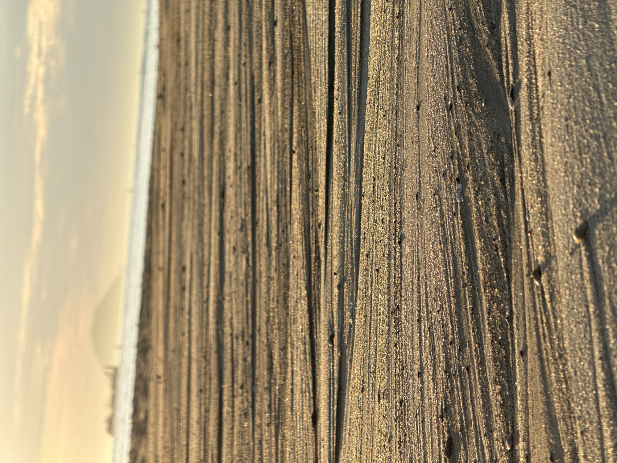 This low-angle photograph captures a sandy beach scene at either sunrise or sunset in Đà Nẵng, Vietnam. The foreground is dominated by wet sand exhibiting numerous linear ripple patterns and small, dark pebbles, intensely illuminated by a warm, golden light that casts distinct, elongated shadows to the left. The midground comprises a blurred expanse of the ocean. The distant background, also significantly blurred, features a hazy sky displaying pale yellow, orange, and light blue tones. On the far horizon, a large, dark ship is silhouetted against the lighter sky on the left. To its right, a prominent, rounded mountain or peninsula, consistent with Son Tra Peninsula, rises from the horizon. No individuals or direct interactions are visible.