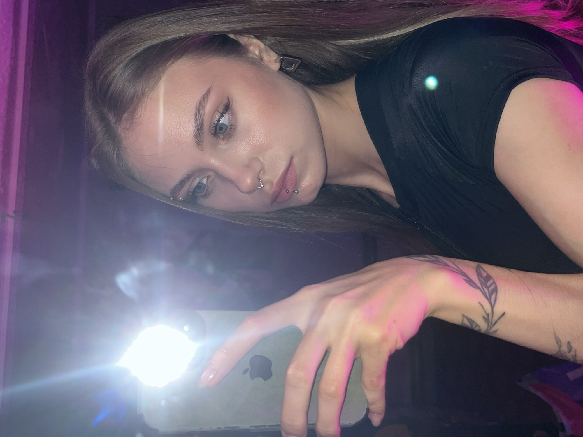 A young adult female is holding a smartphone with its flashlight on, directed towards the viewer. The light from the flash dominates the foreground. The female has light skin, light brown hair, and multiple piercings, including a nose ring and a lip piercing. She is wearing a black top. A tattoo of a plant with leaves is visible on her left wrist. The background is dark with some hints of purple lighting. The location is not discernible and cannot be determined to be in the city of Фоки, Russia based on the image.