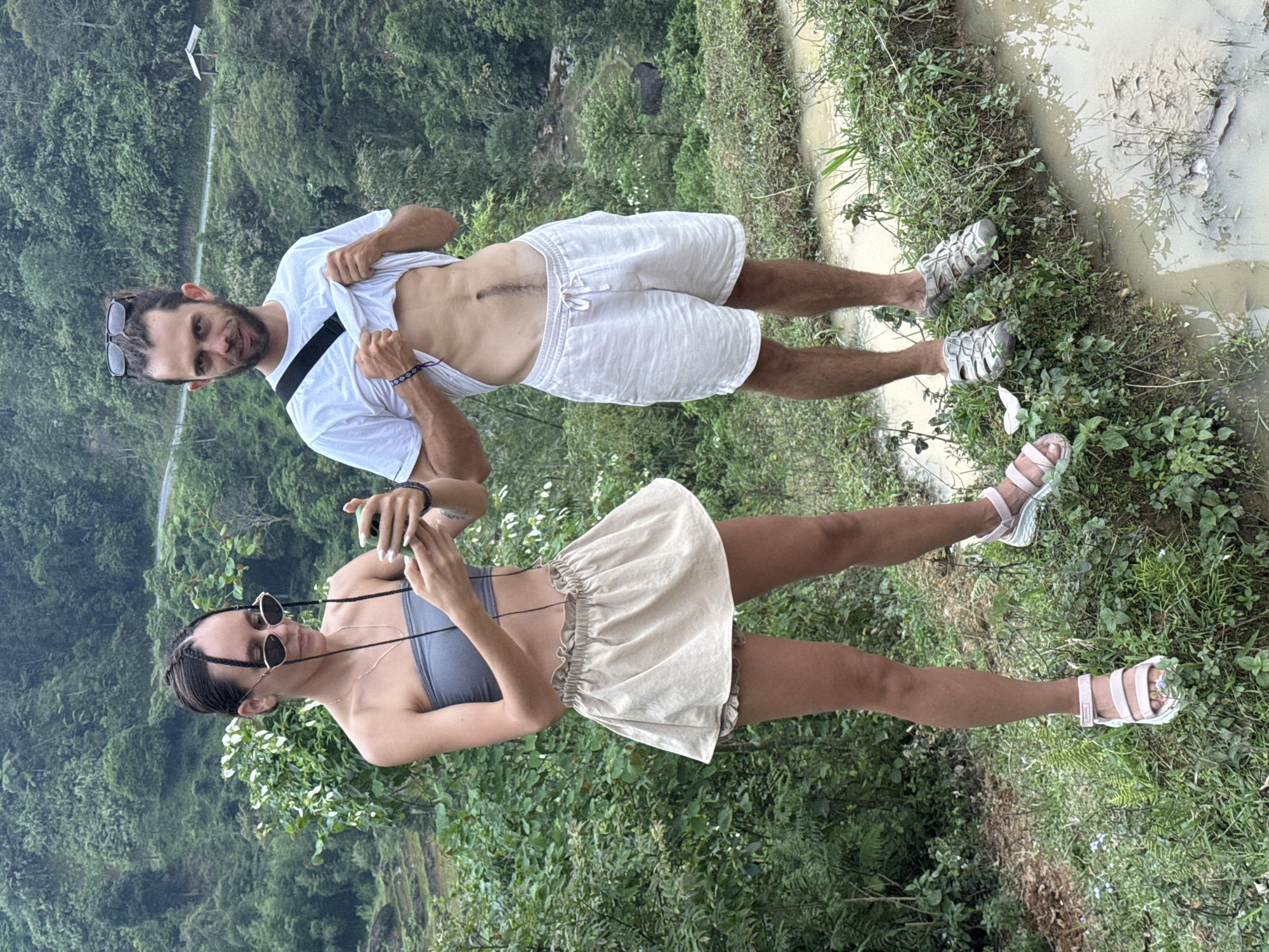 The image captures a man and a woman standing outdoors in a lush, mountainous landscape within Nậm Dịch Commune, Vietnam.
The man, on the right, has a beard, dark hair with some braids, and sunglasses pushed onto his head. He wears a white short-sleeved t-shirt, which he is pulling up with both hands to reveal his lean abdomen, showing a prominent vertical scar and muscle definition. He is also wearing white linen-style shorts and sturdy grey sandals. A black strap, possibly from a cross-body bag, runs across his chest. His feet are positioned near a muddy, water-filled channel bordered by dense green plants.
The woman, on the left, has dark braided hair and is wearing round sunglasses. She is dressed in a grey strapless crop top and a light beige, ruffled mini-skirt. She holds a smartphone with both hands, appearing to be interacting with it or taking a photo. Her legs are bare, and she wears light pink and white sandals, also standing near the edge of the vegetation.
The ground around them is a mix of dirt, green plants, and muddy water, suggesting a natural and possibly damp environment. In the background, rolling hills or mountains are covered in dense, vibrant green foliage, indicating a heavily forested area. A narrow road or path can be faintly seen winding through the distant slopes. The lighting appears soft and diffused, typical of an overcast day. No visible text is present in the image. The overall scene suggests the couple is exploring a scenic, rural area, possibly on a trek or outing.
