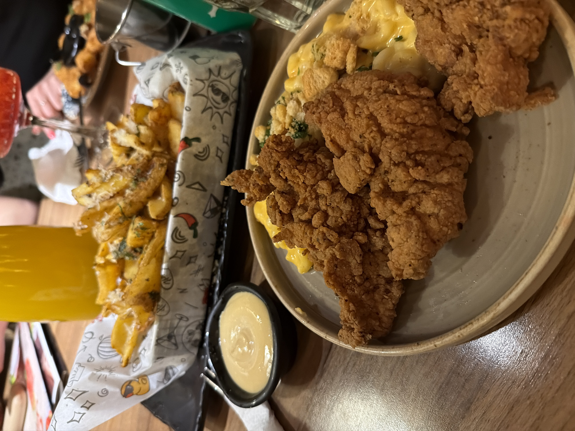 A close-up, slightly angled overhead shot shows a dining table with multiple dishes of food. On the right side of the image is a plate holding three pieces of fried chicken and a portion of mac and cheese topped with croutons and herbs. To the left, a basket lined with patterned paper contains French fries topped with seasonings. A small black bowl holds a dipping sauce. In the background, obscured by the main dishes, are additional food items. A glass of orange juice is visible. The setting seems to be a restaurant, possibly in Sofia, Bulgaria, but there's no definitive evidence within the frame. No people or specific interactions are distinctly visible.